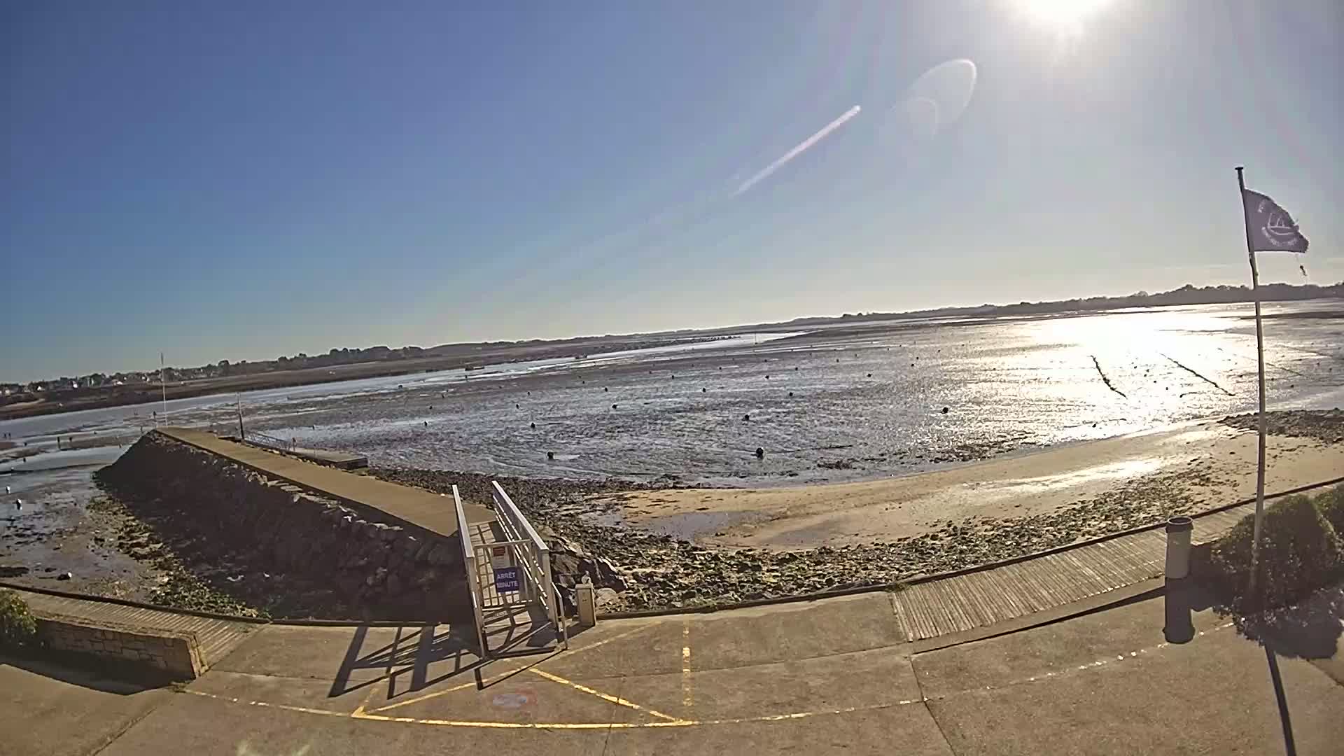 A sunny day reveals a low tide, exposing a wide expanse of mudflats with scattered buoys and a stone jetty leading to a sandy beach.