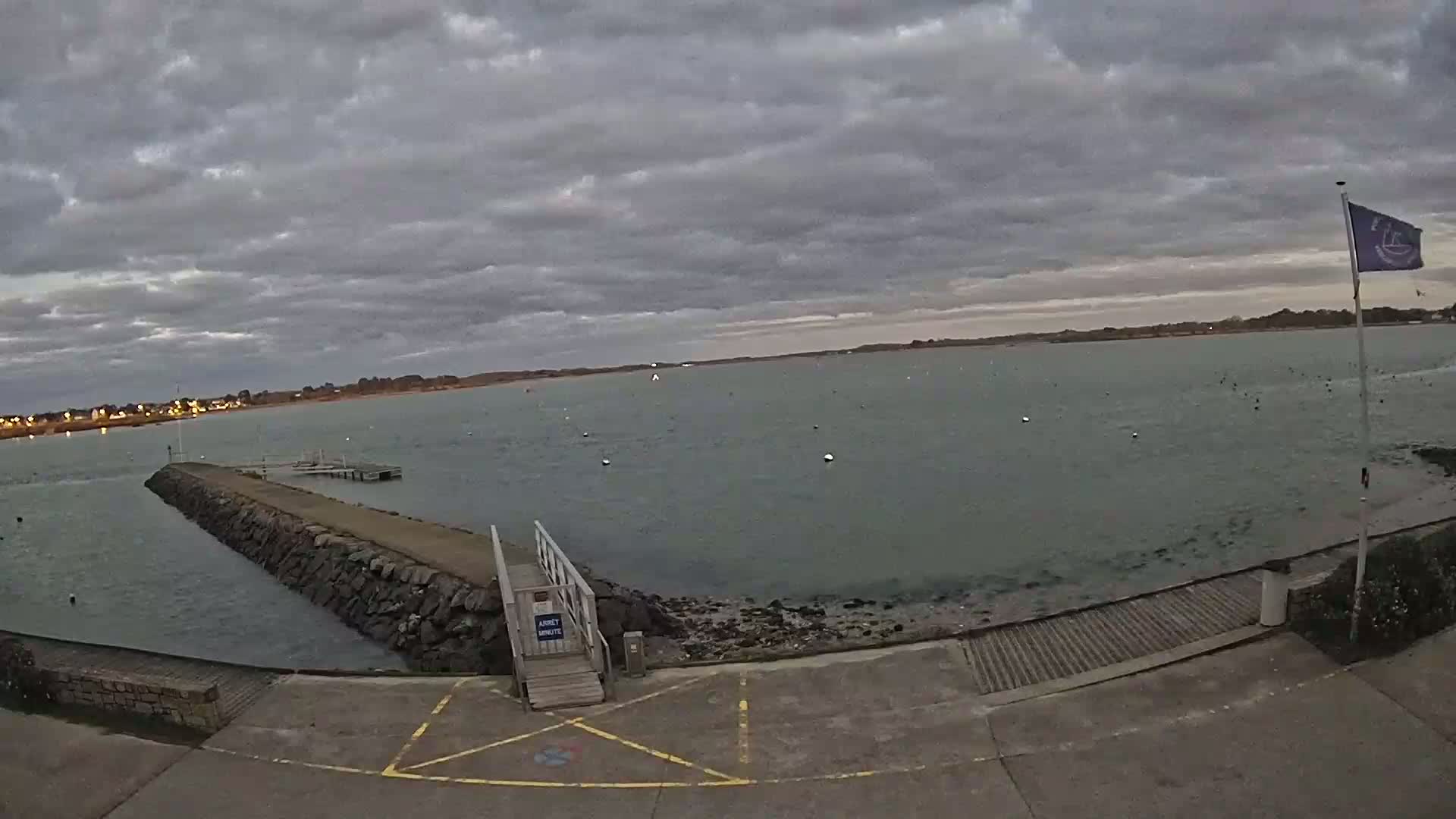 A calm body of water is seen under a cloudy sky, with a stone jetty extending into it and a small ramp leading to the water's edge.