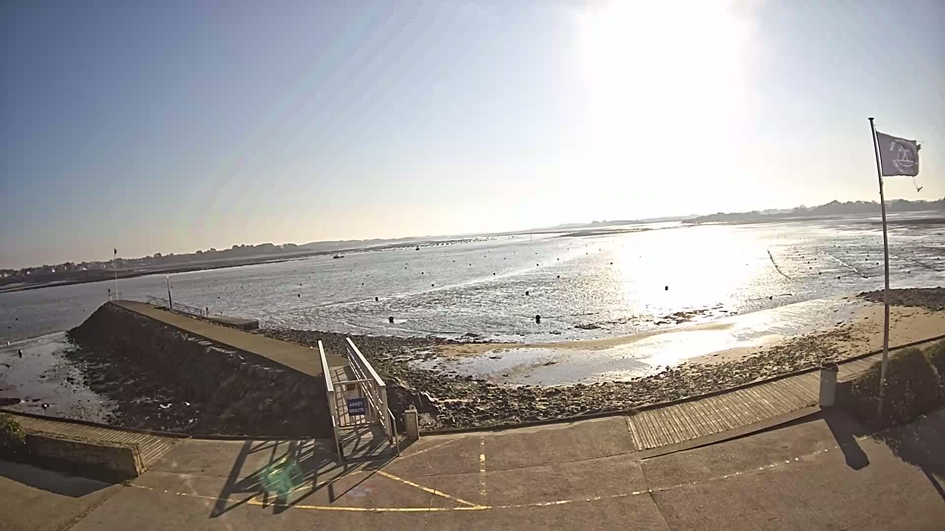 A sunny day reveals a calm body of water with a low tide exposing a muddy shoreline, a stone jetty with a gate, and a flagpole with a flag on a paved area.