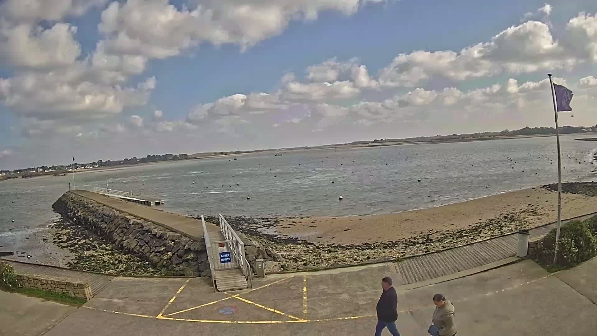 A rocky jetty extends into a calm body of water under a partly cloudy sky, with two people walking on a paved area nearby.