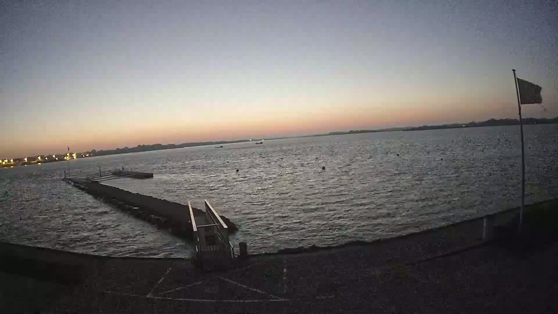 A calm body of water at sunset, with a small pier extending from the shore, and a flagpole with a flag on it.