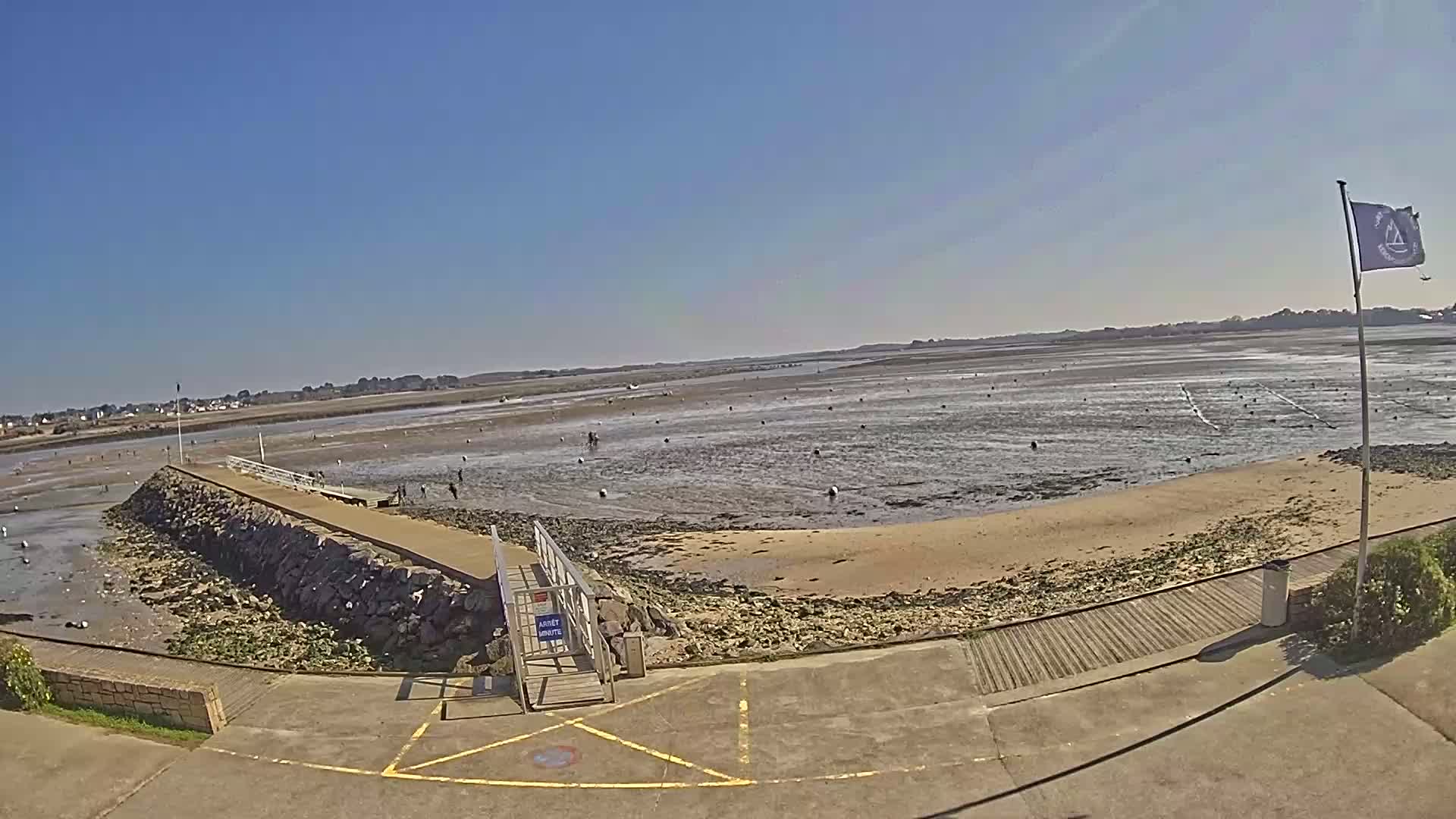 A stone pier extends into a wide, muddy intertidal zone under a clear blue sky, with a sandy beach and several people visible in the distance.