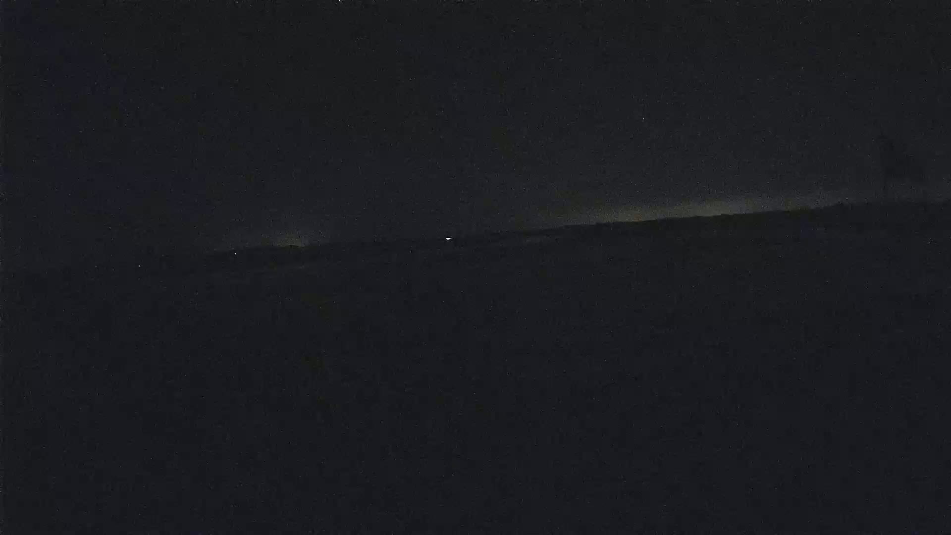 A dark, nighttime landscape shows a flat horizon line with a few faint lights in the distance under a mostly clear sky.