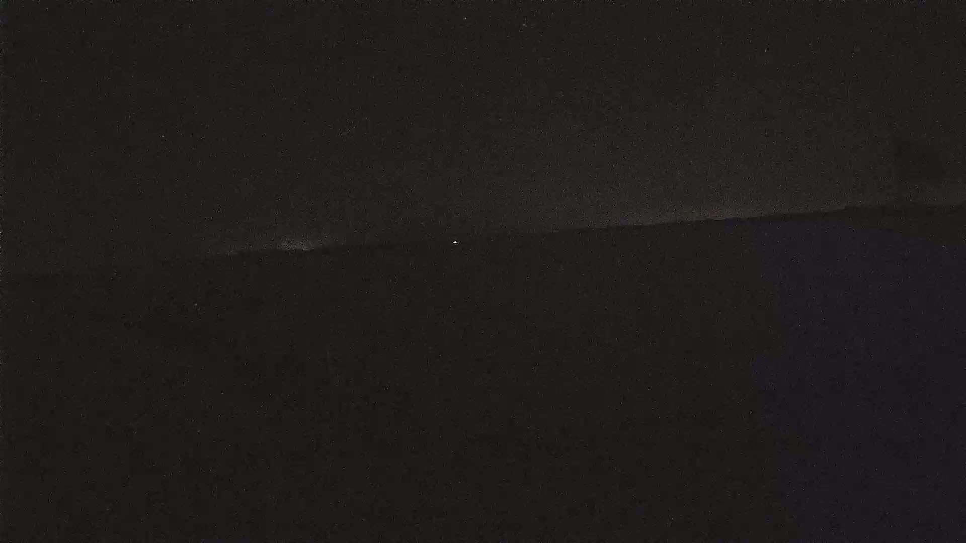 A dark, nighttime landscape is visible with a single faint light in the distance under a mostly clear sky.