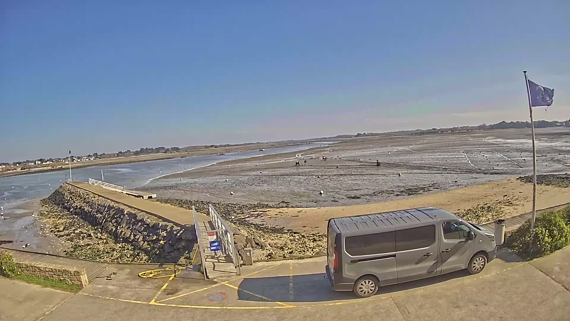 A gray minivan is parked near a stone jetty overlooking a wide expanse of exposed mudflats and calm water under a clear blue sky.
