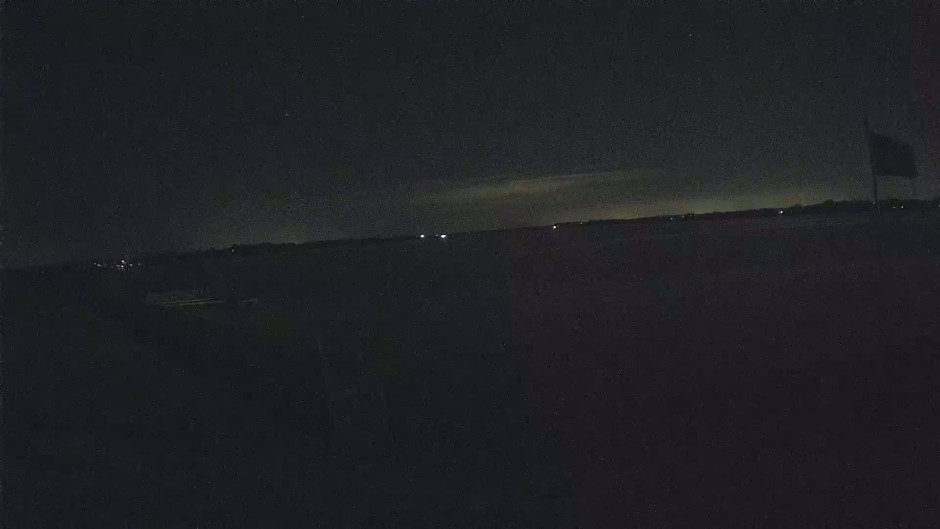 A dark, nighttime landscape shows a flat horizon line with a few distant lights under a mostly clear sky.