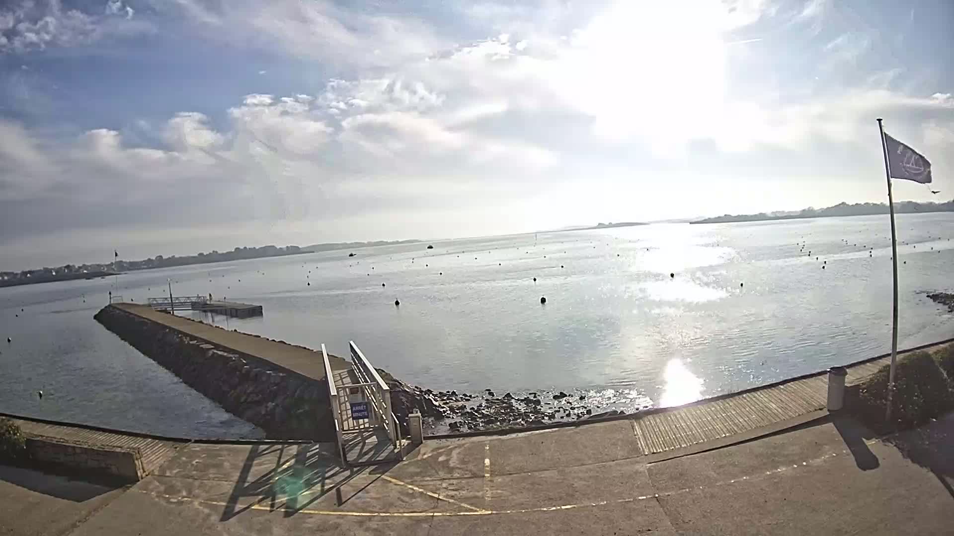 A calm, sunny day reveals a harbor scene with a long jetty extending into the water, numerous buoys scattered across the bay, and a flagpole standing on the shore.