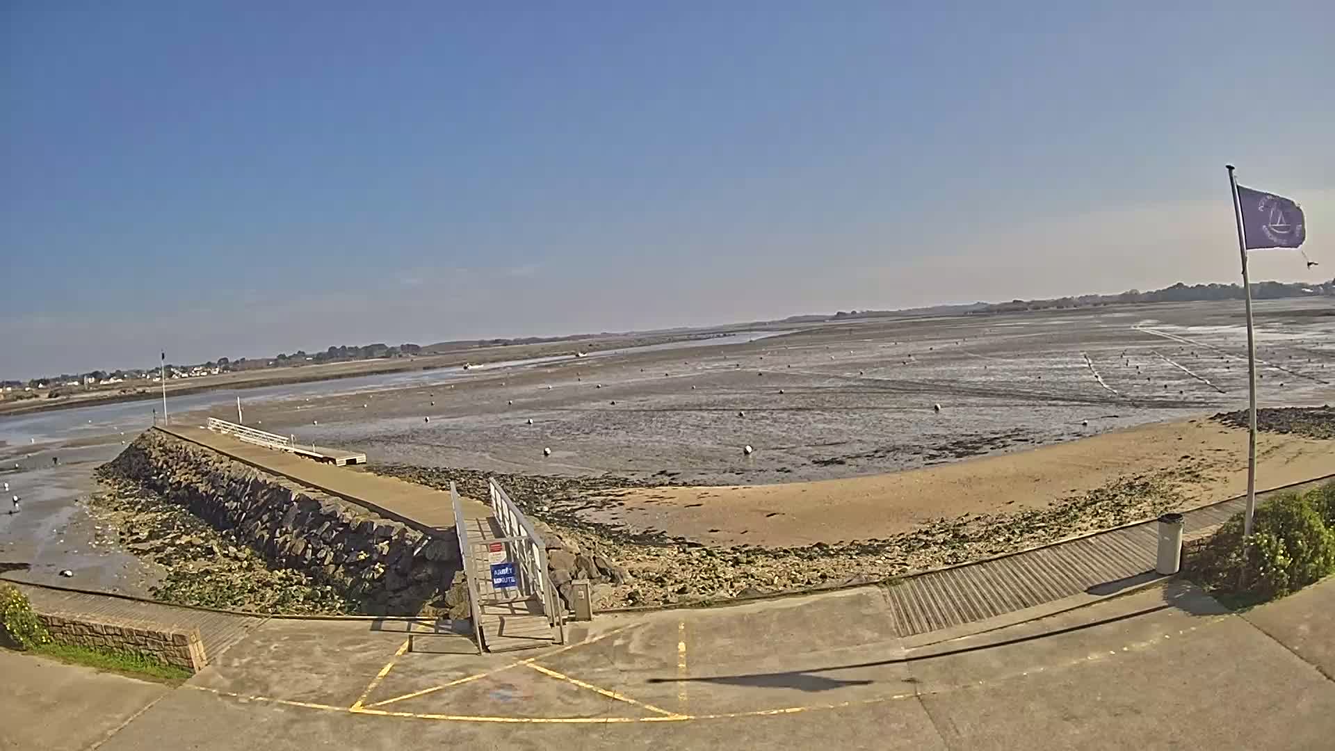 A stone pier extends into a wide expanse of exposed mudflats under a clear blue sky, with a sandy beach bordering the water.
