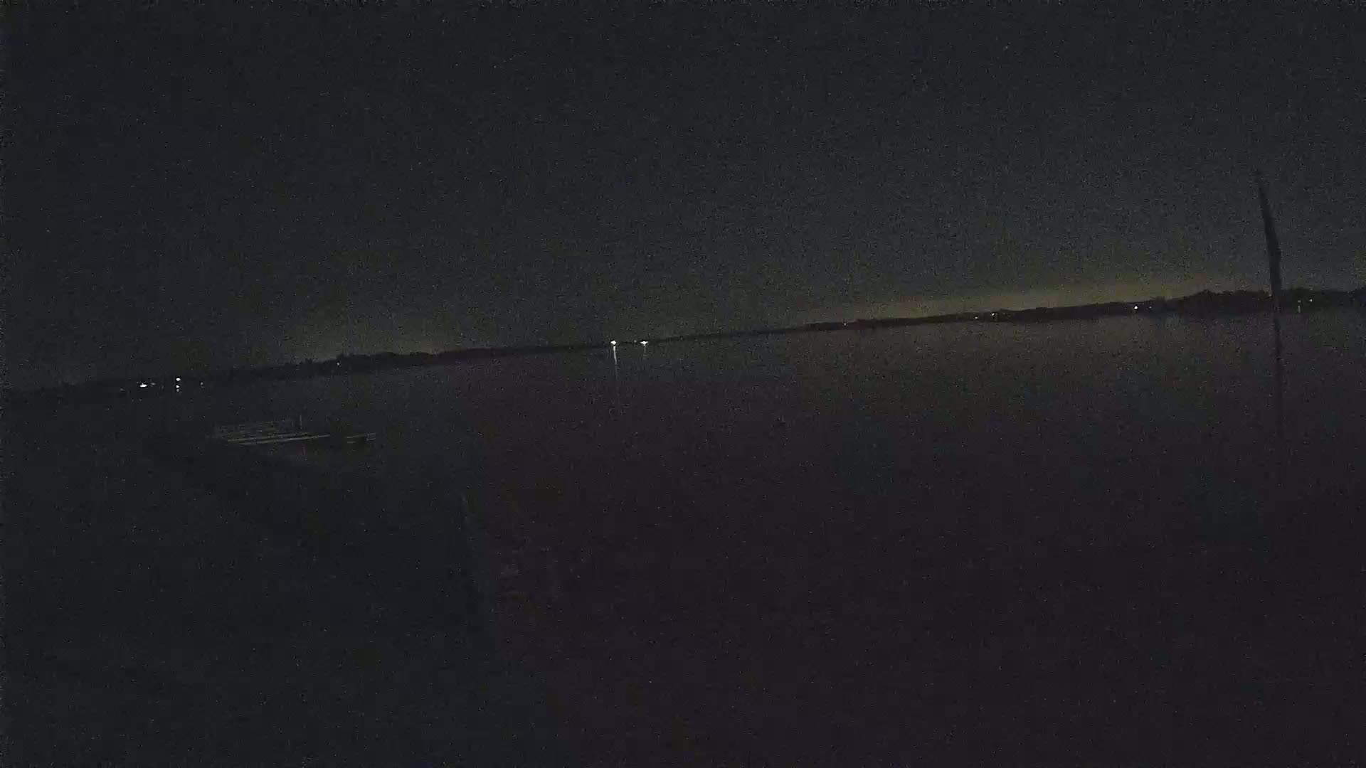 A dark, nighttime view of a calm body of water with a distant shoreline and faint lights visible across the water.