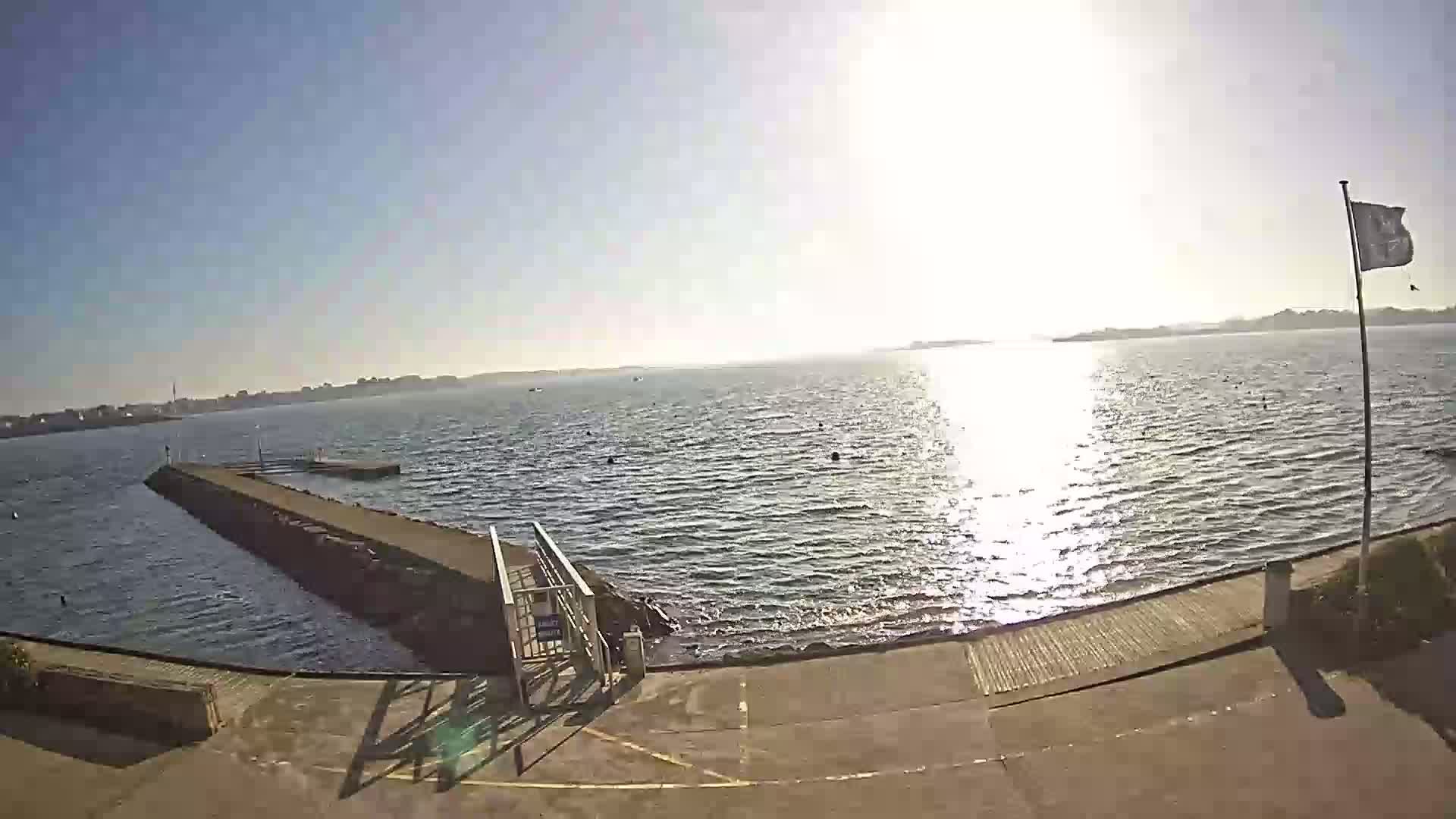 A sunny day at a waterfront location with a small pier extending into a calm body of water, a flagpole with a flag visible in the background.