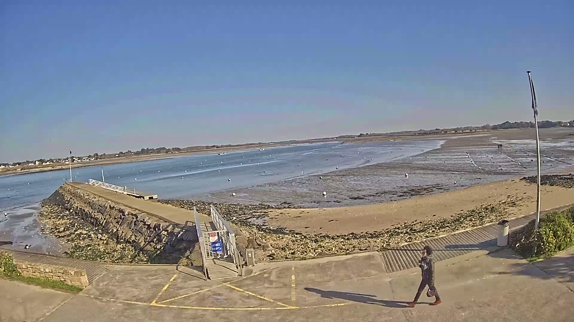 A person walks along a paved area near a stone jetty extending into a calm, shallow body of water under a clear blue sky.
