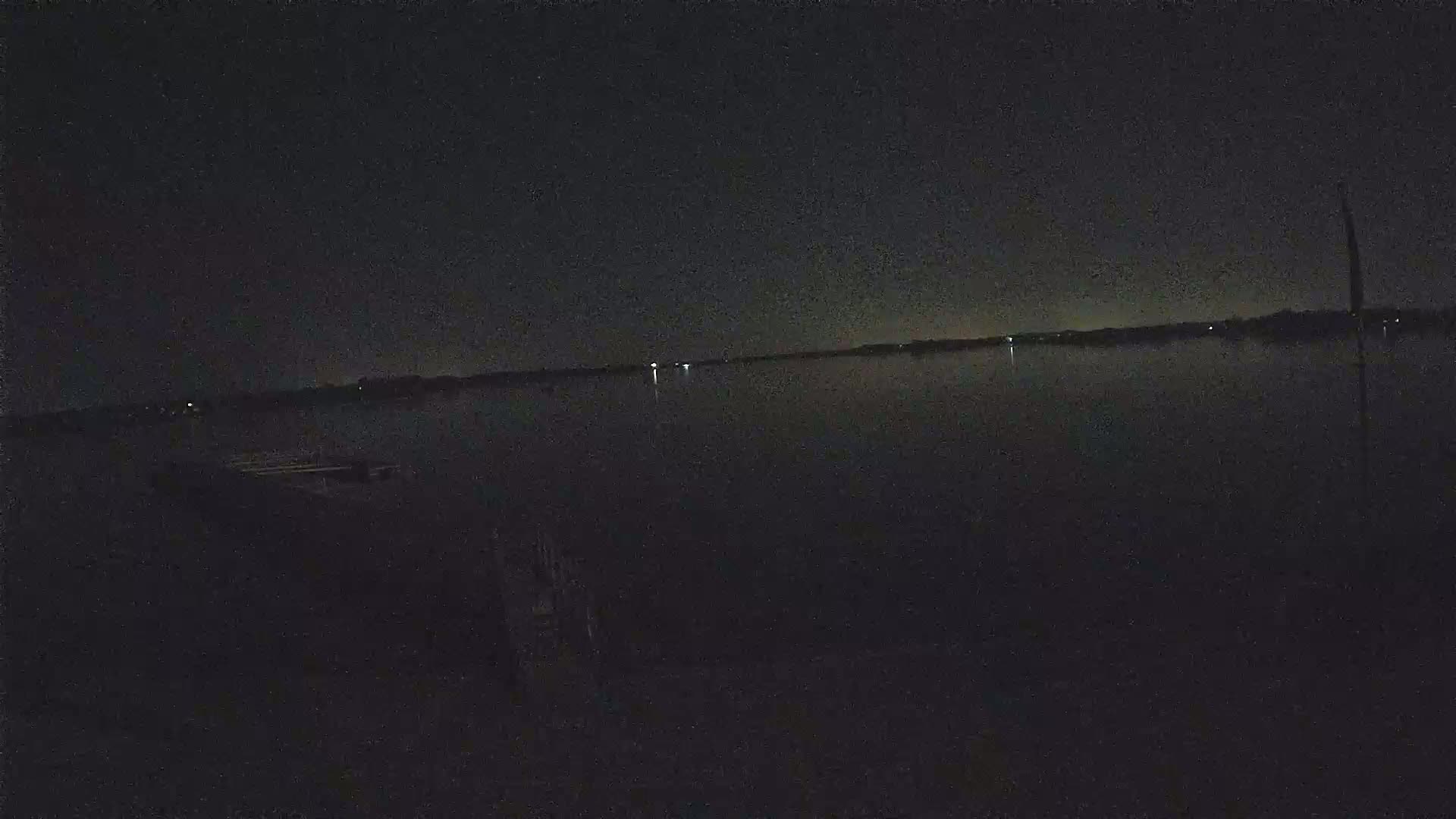 A dark, calm body of water is seen at night, with a distant shoreline barely visible under a mostly clear sky.
