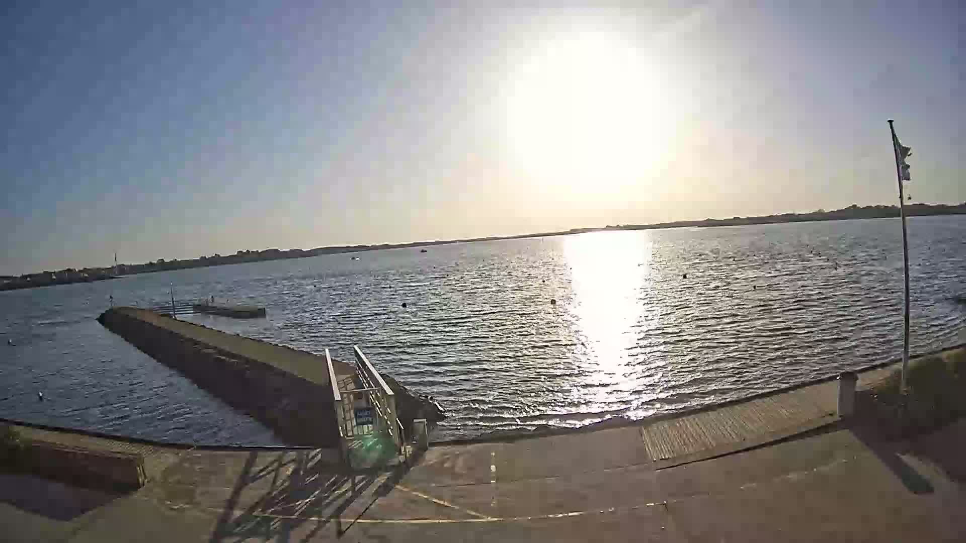 A calm body of water under a bright, sunny sky is viewed from a boat launch with a small pier extending into the water.