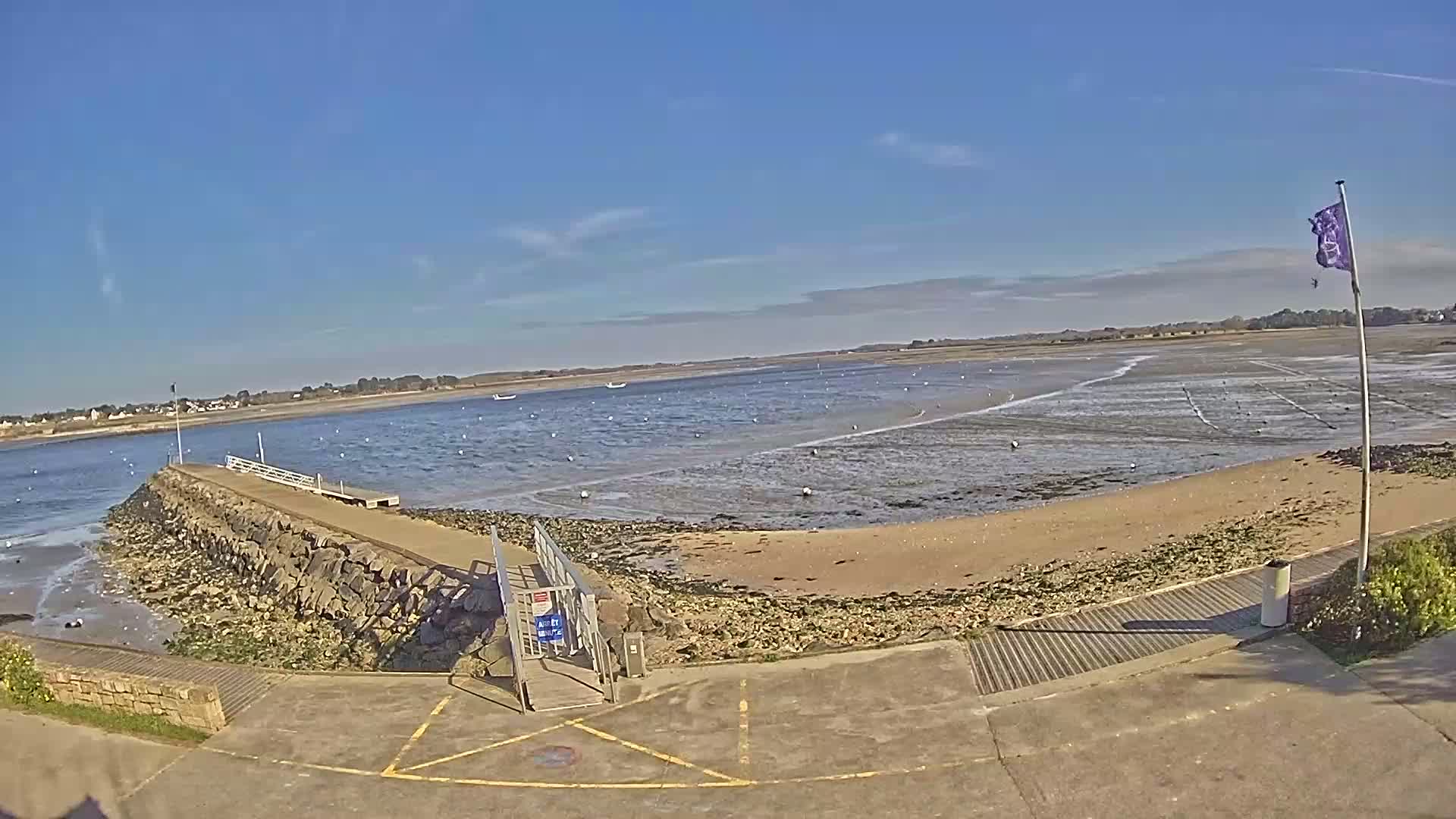 A stone jetty extends into a calm, shallow bay on a sunny day, with a sandy beach visible at low tide.