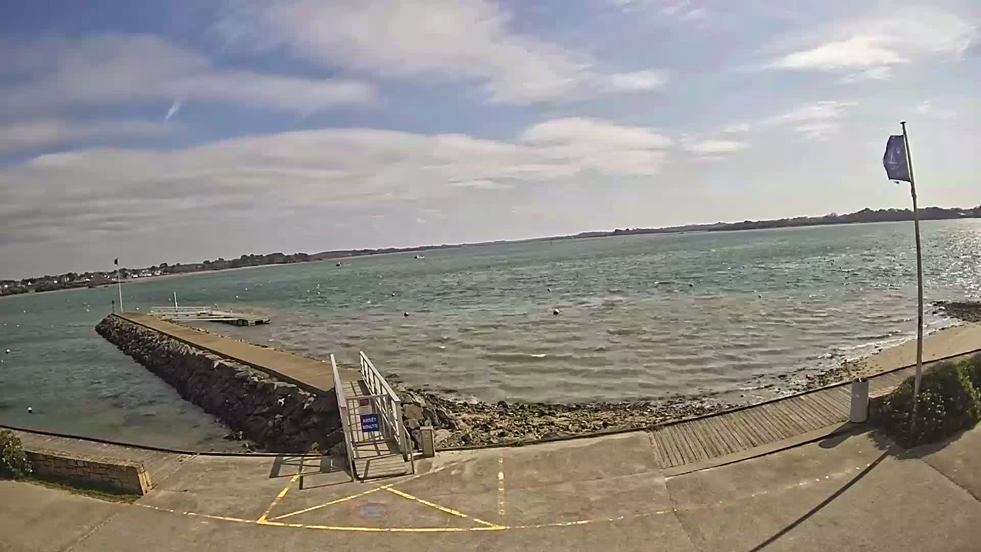 A stone jetty extends into a calm, light-brown body of water under a partly cloudy blue sky.