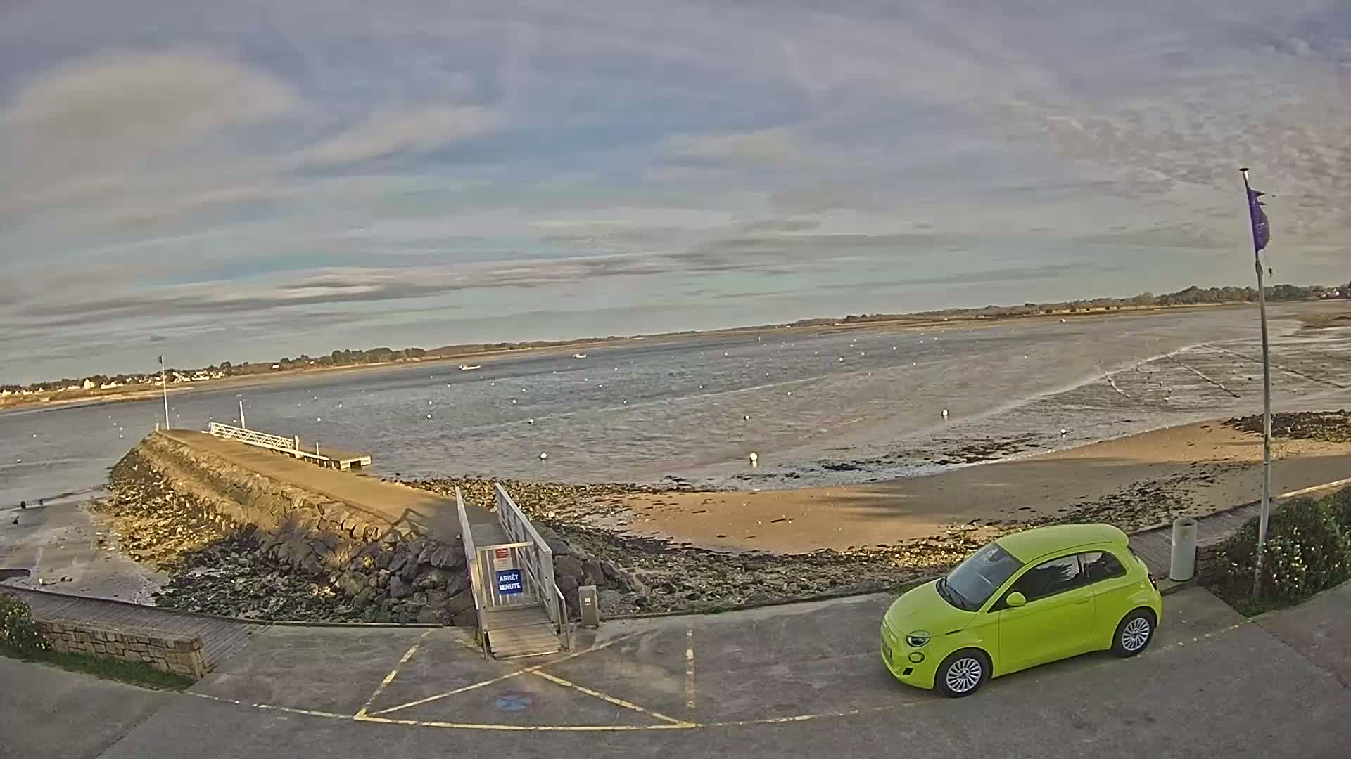 A lime-green car is parked near a rocky jetty extending into a calm, wide body of water under a mostly cloudy sky.