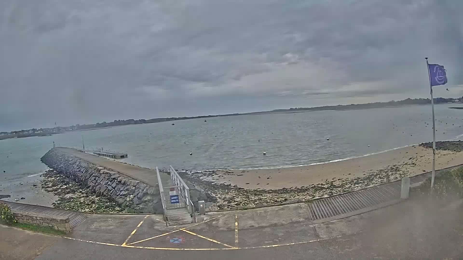 A stone jetty leads to a small sandy beach on a calm, overcast day, with a body of water extending to a distant shoreline.