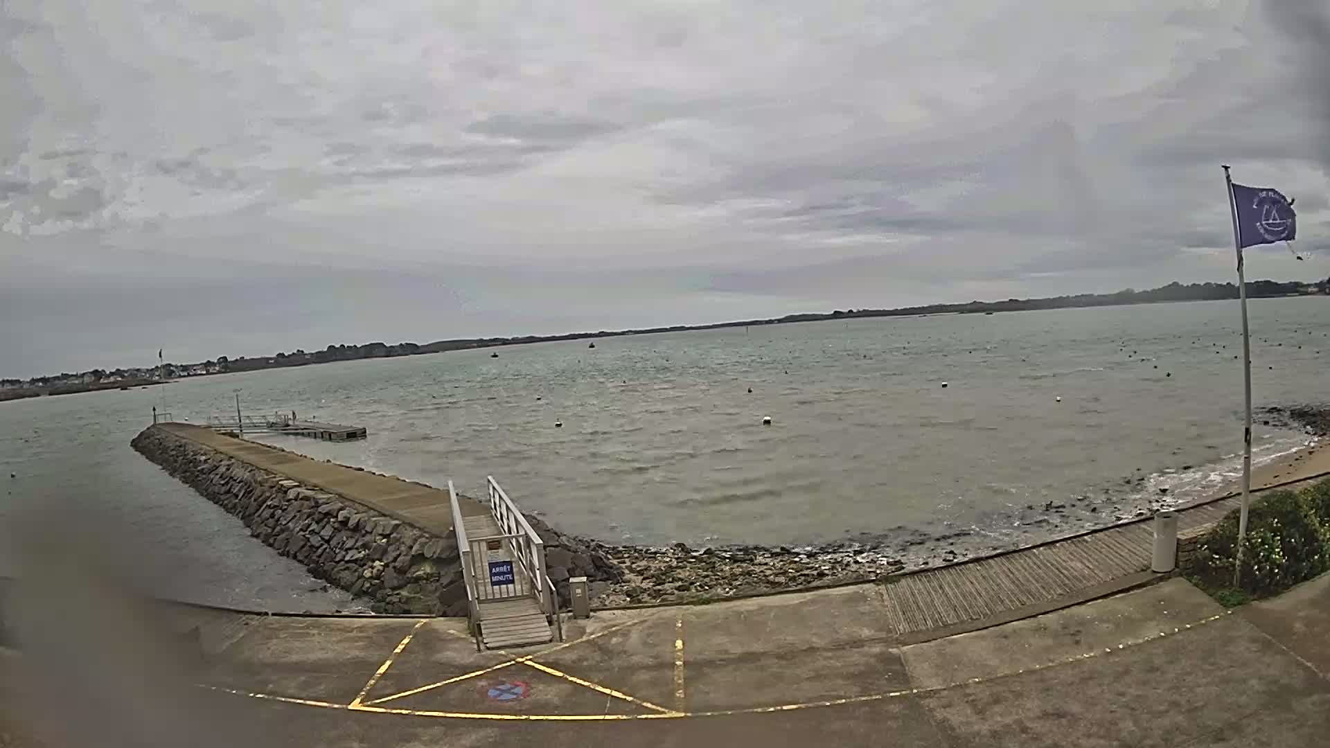 A calm, grey day reveals a stone jetty leading to a small dock extending into a body of water, with a shoreline visible in the background.