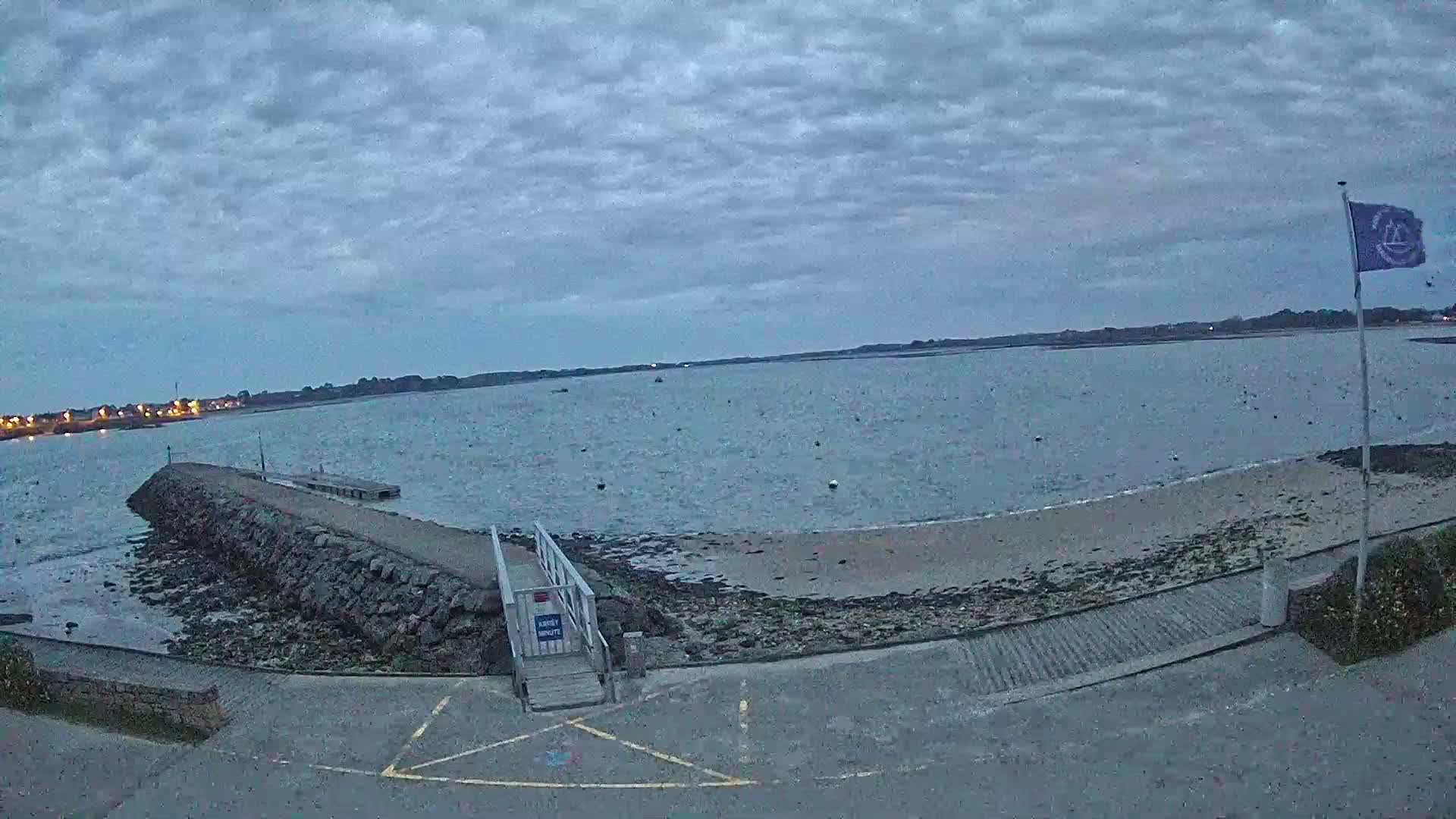 A calm body of water under a cloudy sky is viewed from a shore with a stone jetty, a small wooden walkway, and a flagpole.