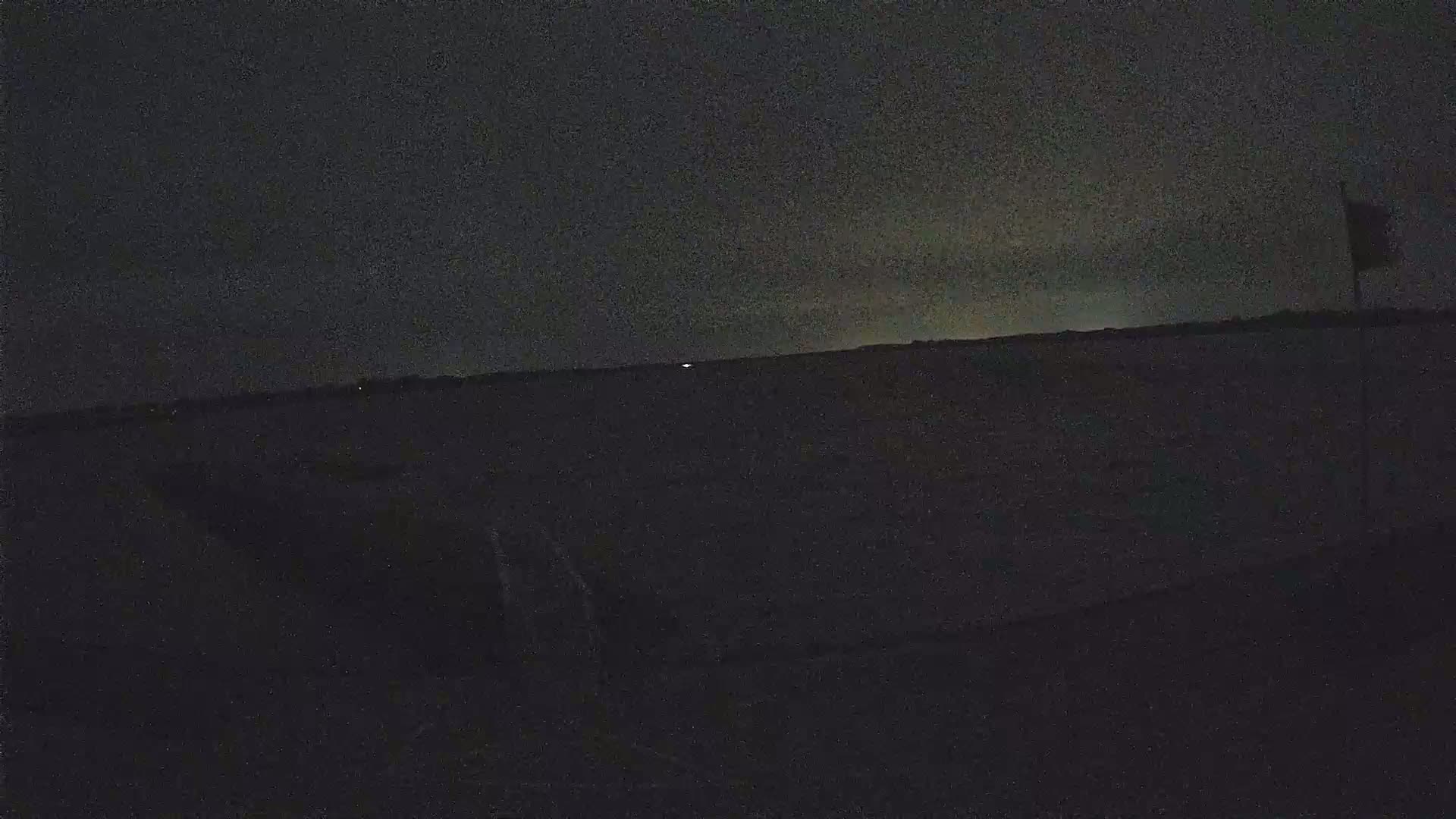 A dark, nighttime scene shows a flat, dark landscape under a mostly clear, dusky sky with a single flagpole visible in the right corner.