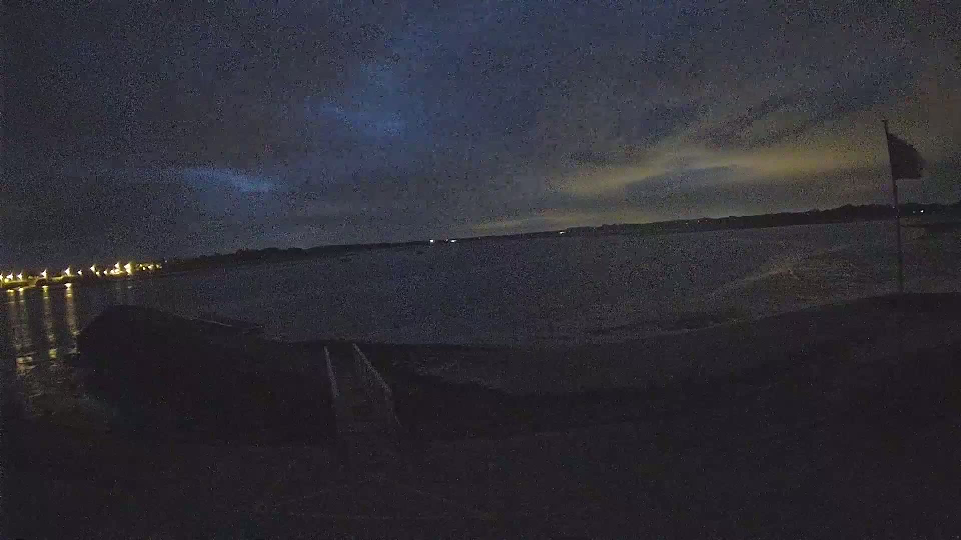 A dark, nighttime scene shows a body of water under a mostly cloudy sky, with a flagpole and a distant shoreline visible.