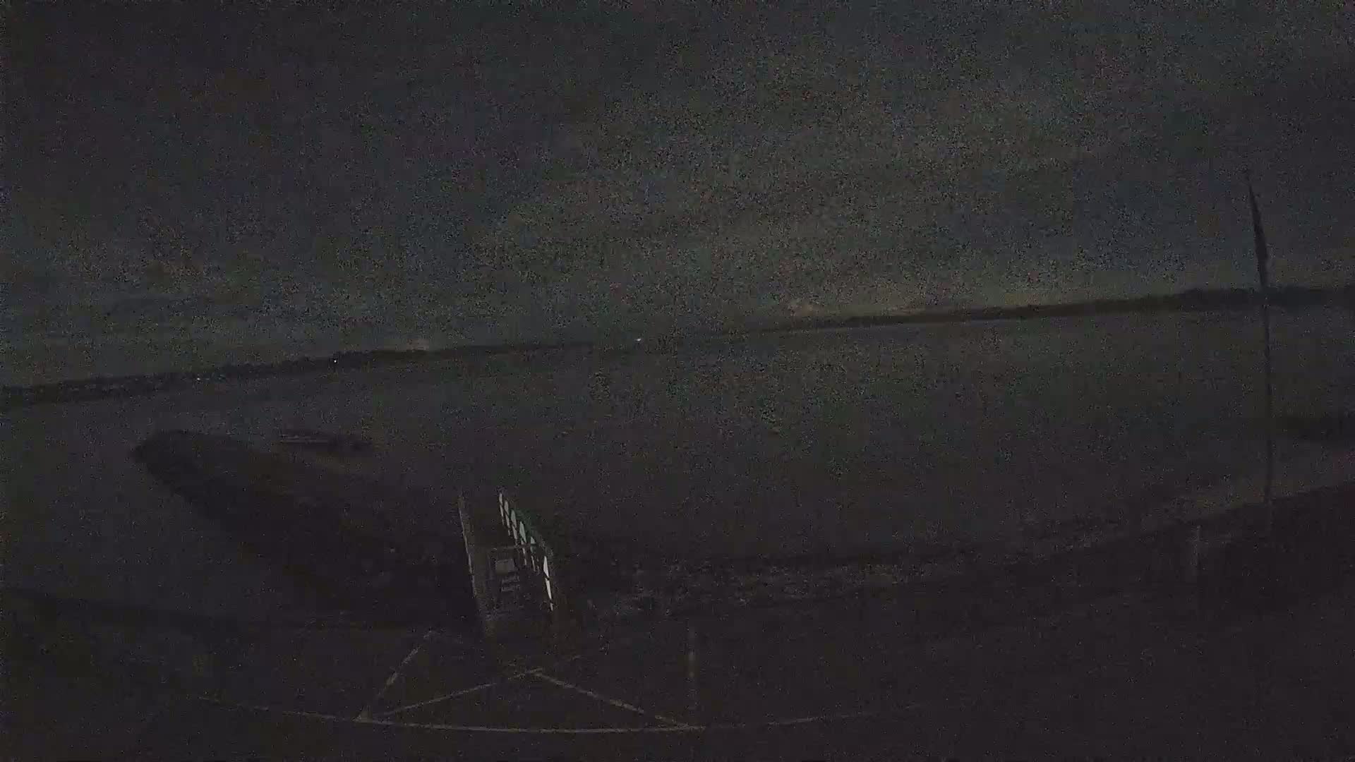 A dark, nighttime view of a calm body of water with a dock in the foreground under a mostly clear sky.