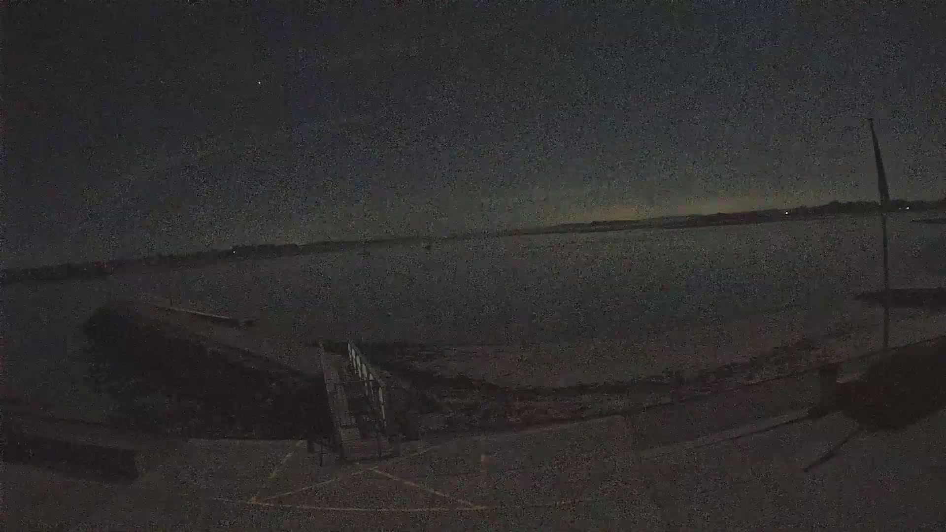 A dark, nighttime scene shows a body of water under a mostly clear sky, with a small dock and a portion of a paved area visible in the foreground.