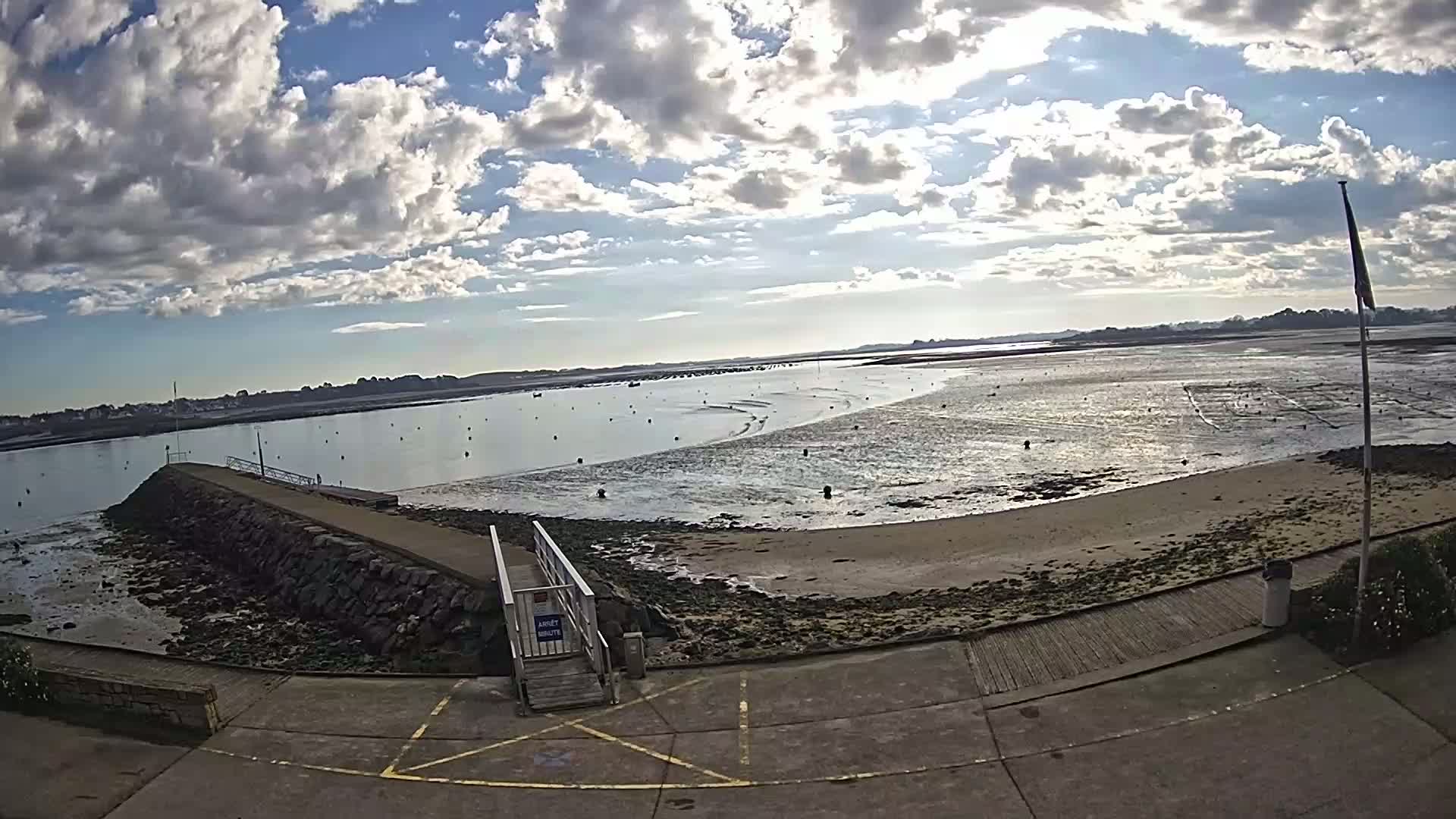 A partly cloudy day reveals a rocky shoreline with a small pier, leading to a sandy beach and calm water dotted with markers, extending to a distant landmass under a partly cloudy sky.