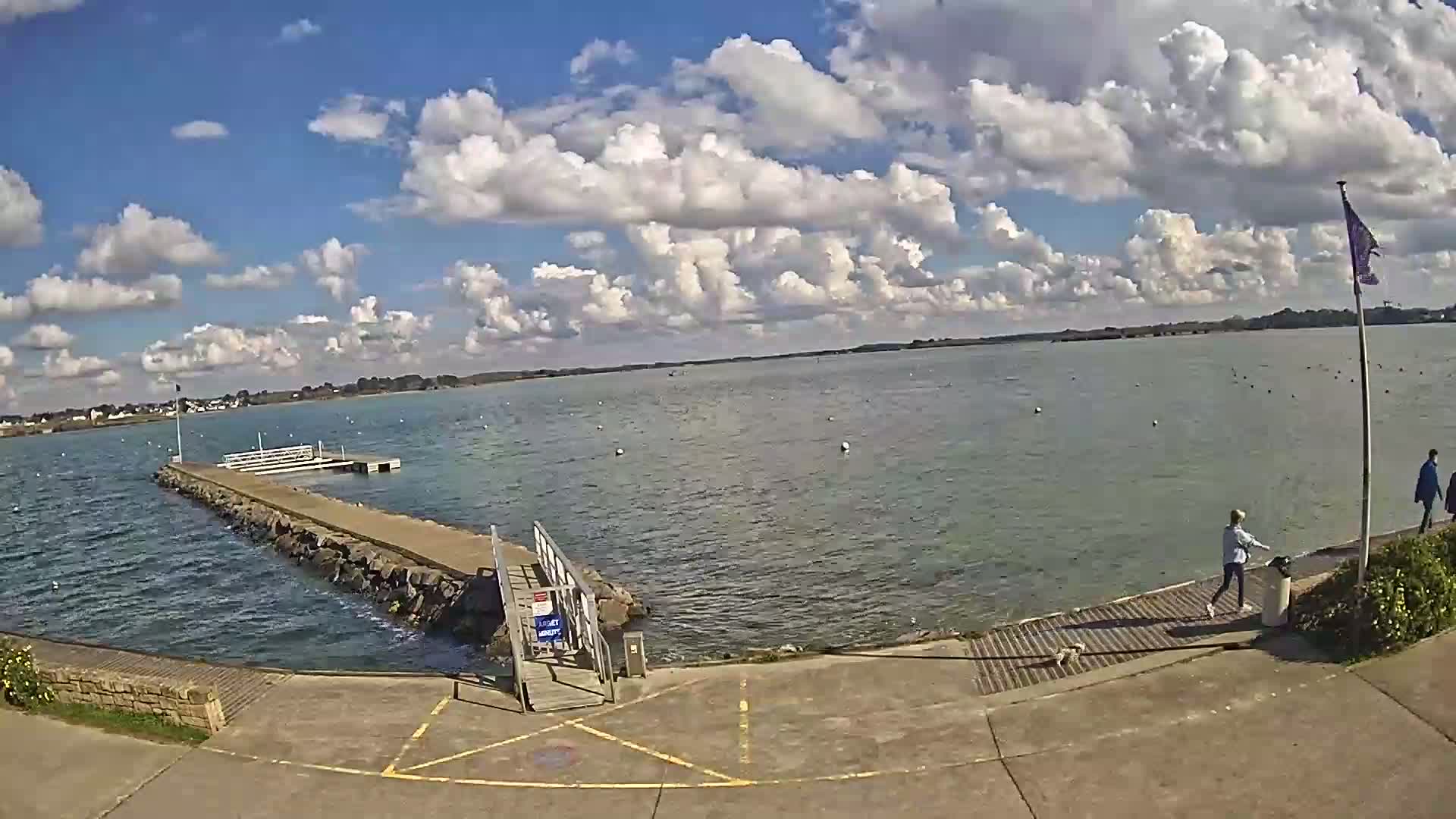 A calm body of water under a partly cloudy sky is viewed from a paved area leading to a small pier.
