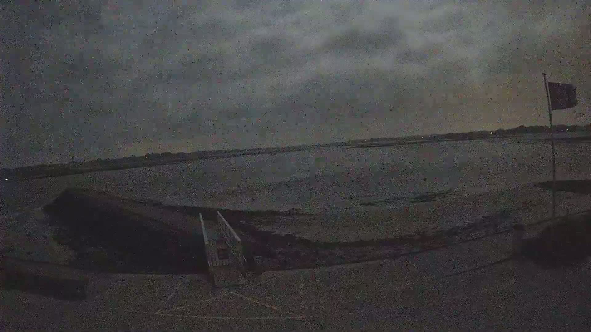 A dark, overcast sky hangs over a calm body of water, a small jetty with a ramp leading to the shore is visible in the foreground, and a flag is partially visible in the distance.