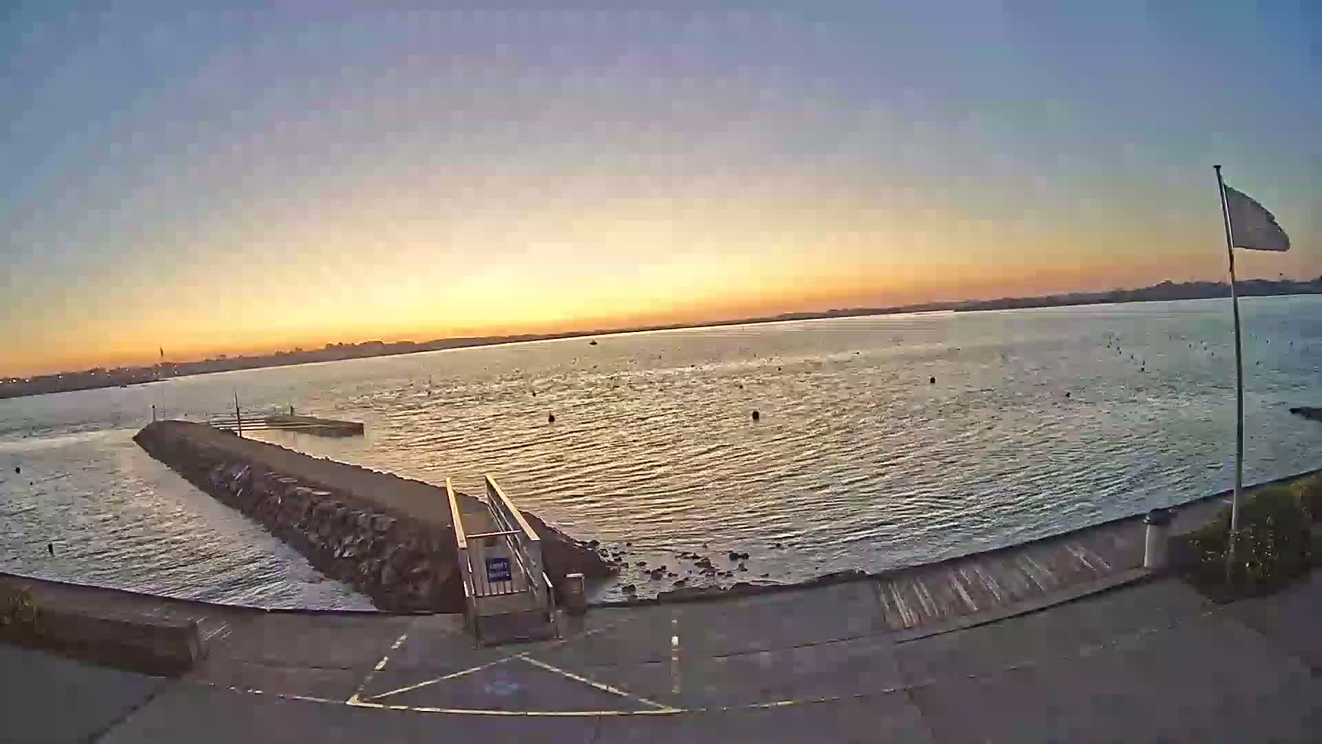 A calm body of water at sunset is viewed from a paved area with a stone jetty and a ramp leading to the water, under a clear sky.