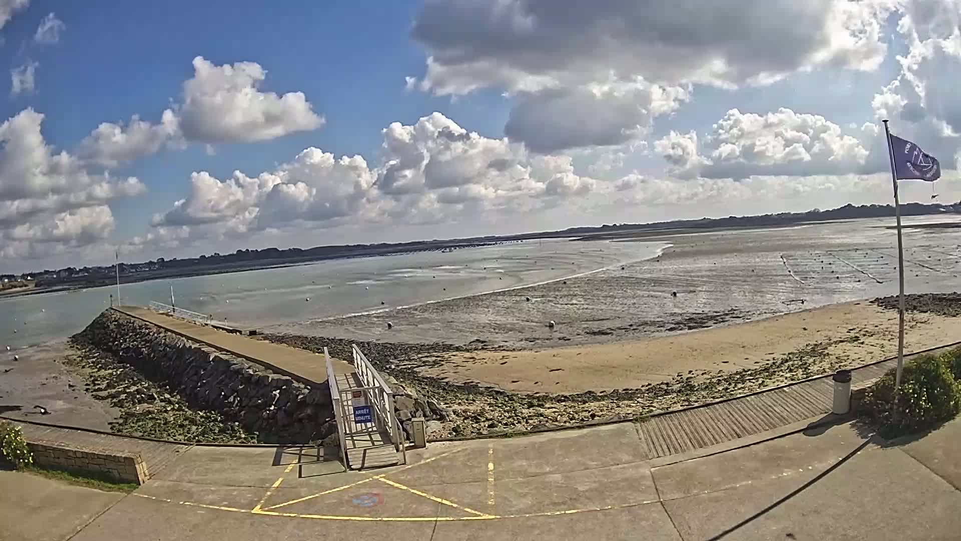 A stone pier extends into a calm body of water under a partly cloudy sky, leading to a sandy beach with exposed mudflats at low tide.
