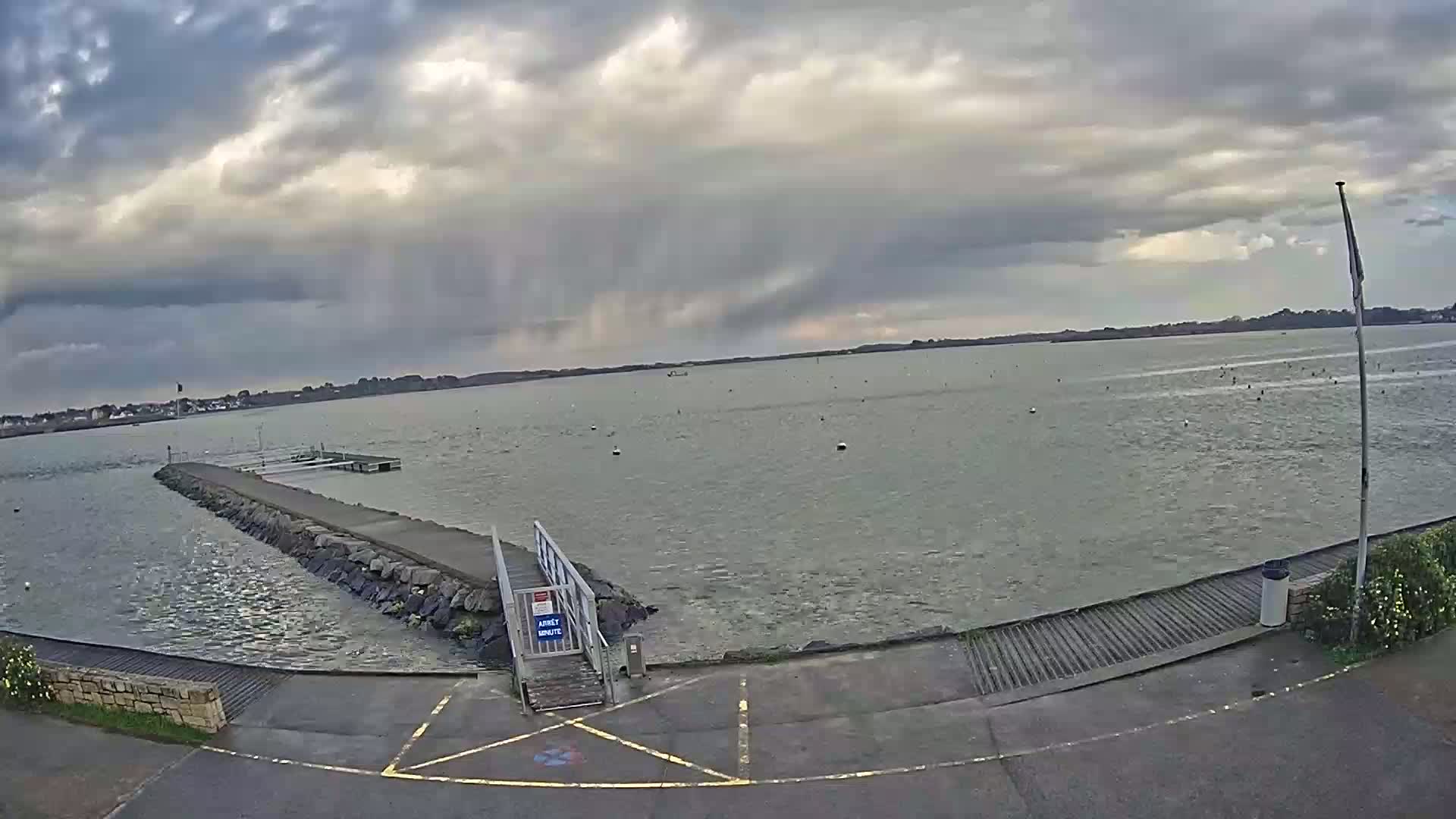 A stone jetty extends into a calm body of water under a cloudy sky with a rain shower in the distance.