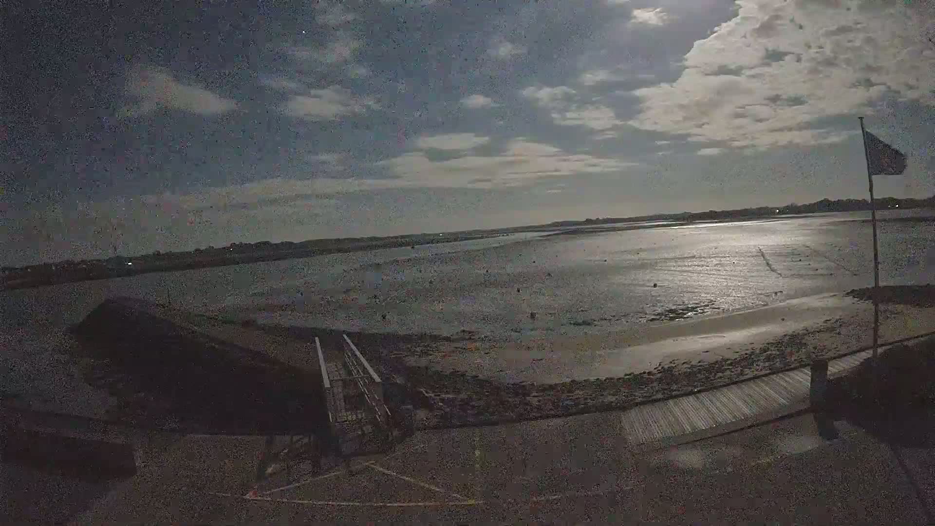 A moonlit night scene shows a calm body of water at low tide, a sandy shore, a walkway, and a flagpole with a flag.