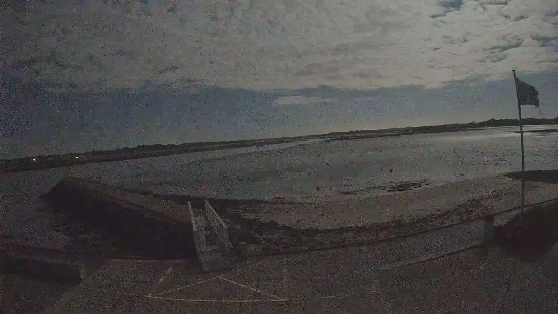 Under a dark, cloudy sky, a low-tide scene shows a wide expanse of mudflats leading to calm water, a small pier, and a flagpole with a flag.