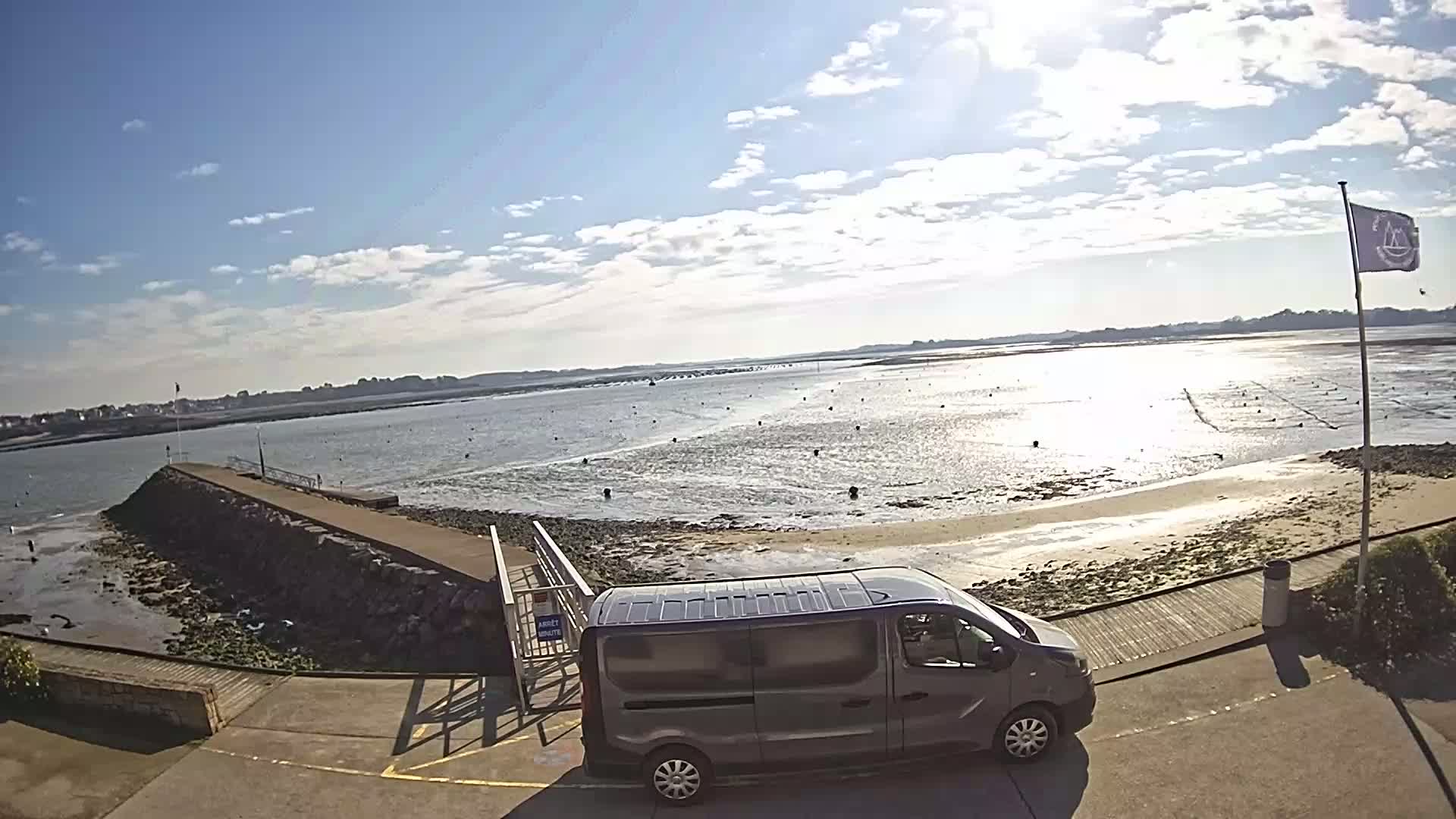 A gray minivan is parked near a stone jetty overlooking a calm, sunlit body of water with a sandy shore and distant oyster beds under a partly cloudy sky.