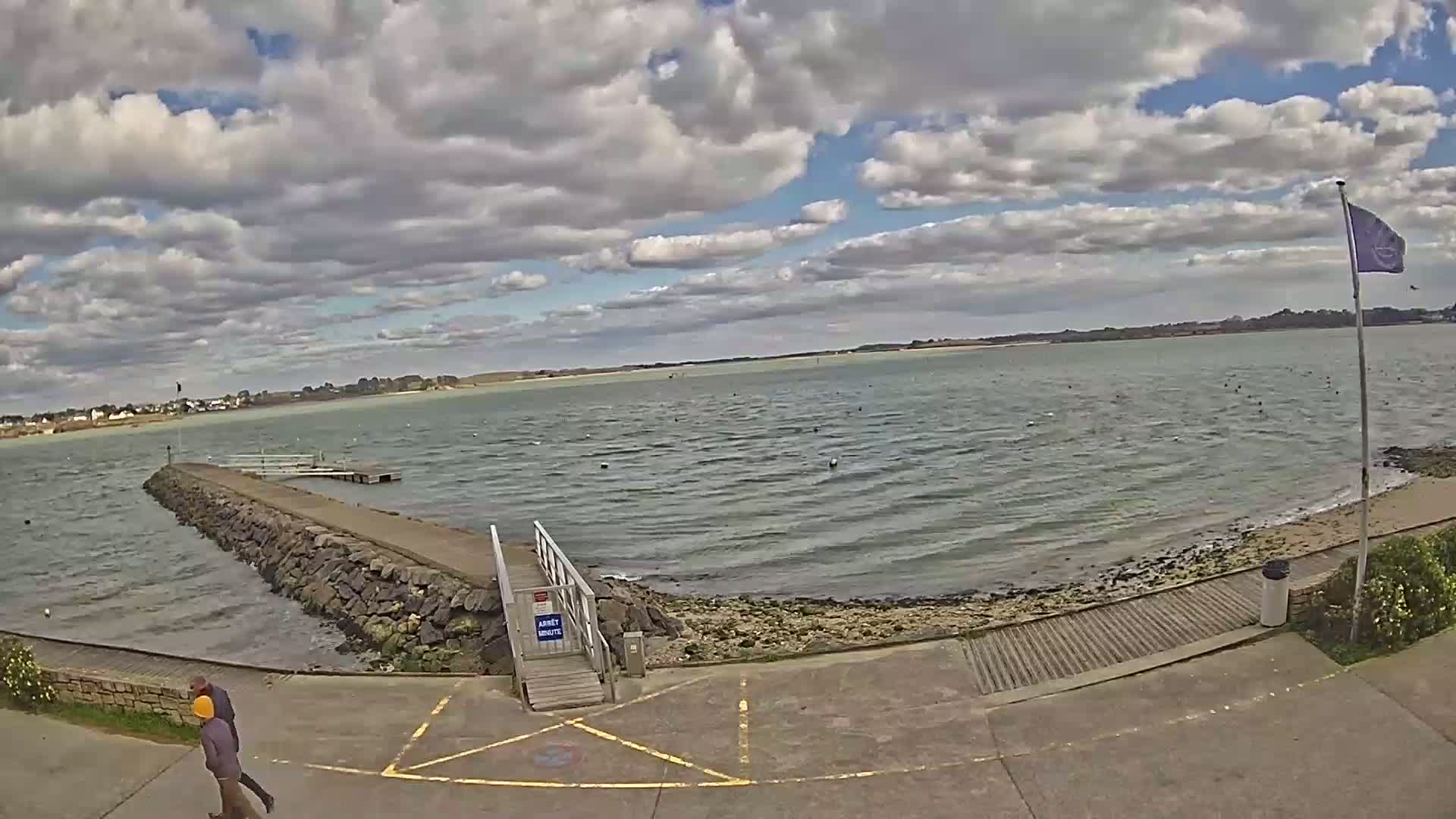 A partly cloudy day overlooks a calm body of water with a stone jetty leading to a small dock, and two people walking near the shore.
