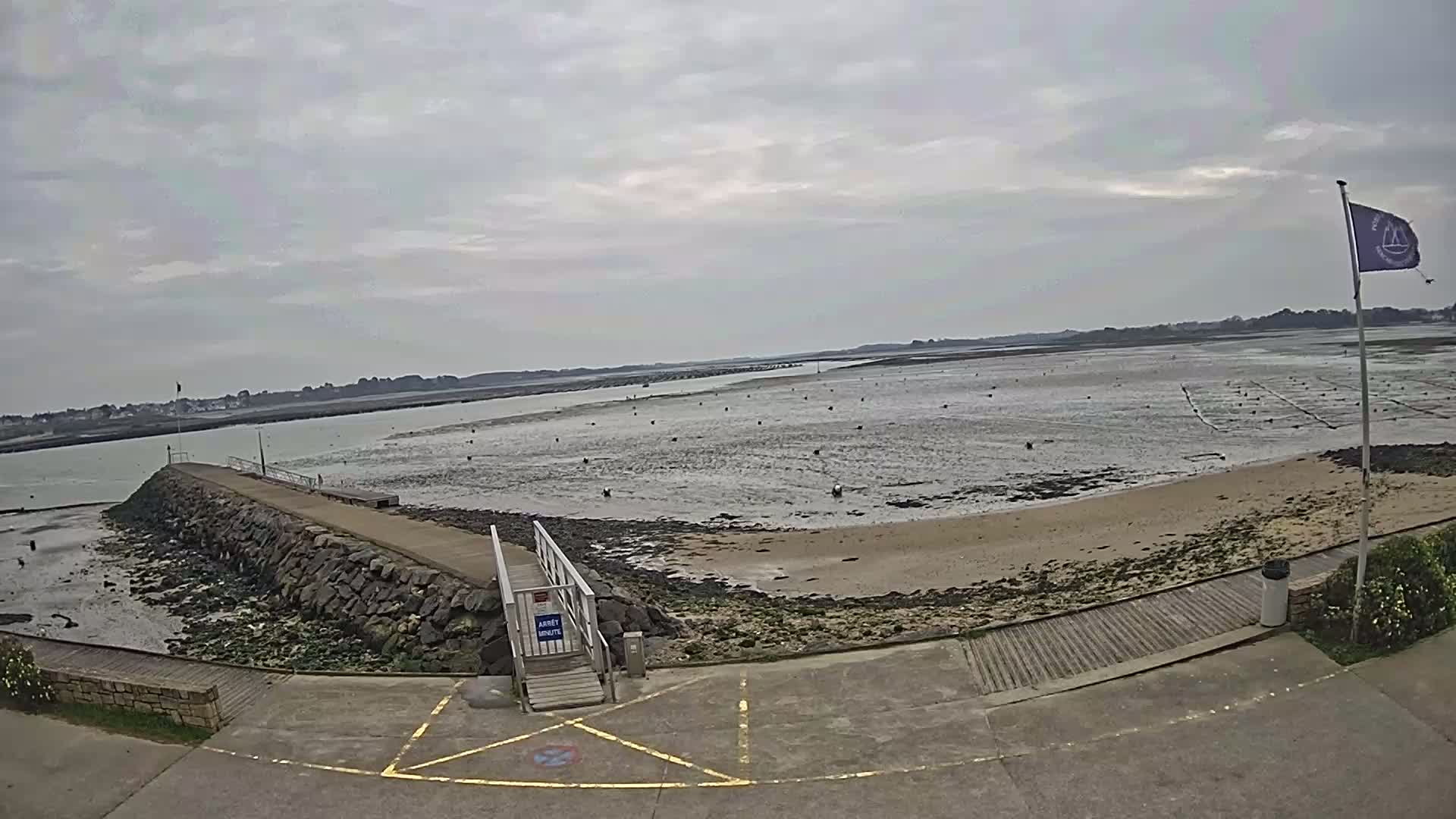 A stone pier extends into a calm, shallow body of water under an overcast sky, leading to a sandy beach with a low tide exposing a grid of shallow water structures.