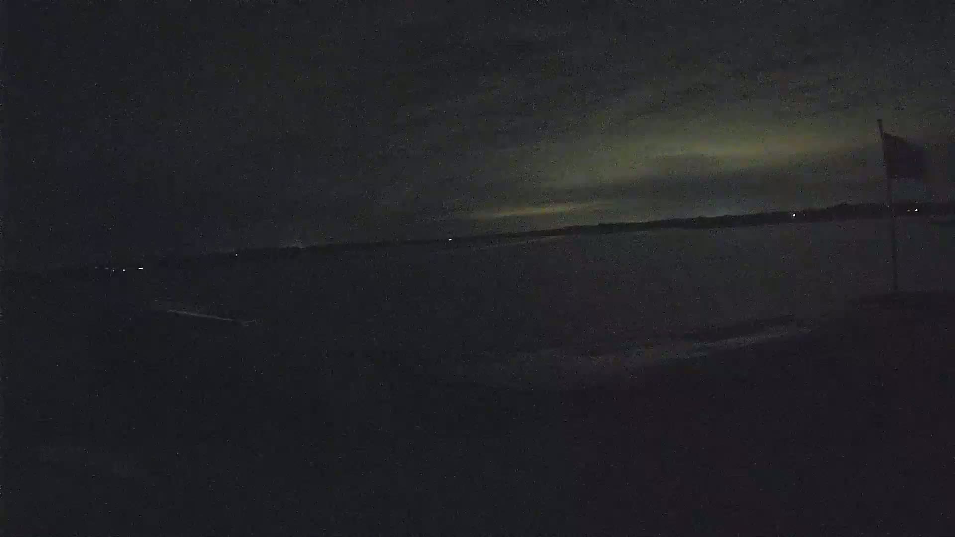 A dark, nighttime scene shows a body of water under a mostly cloudy sky, with a flagpole and flag visible to the right.