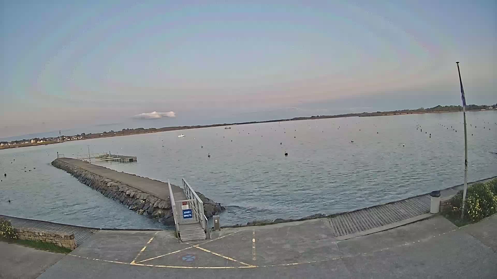A calm body of water with a stone jetty and small boat ramp, under a mostly clear sky.