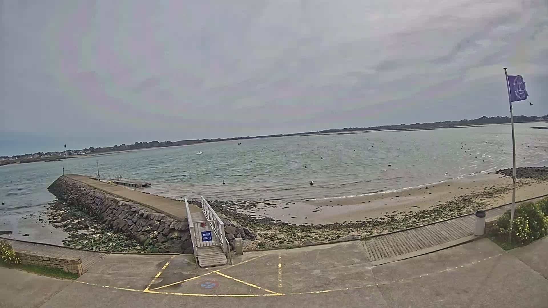 A stone jetty extends into a calm, grey-skied bay with a sandy beach and a few small boats visible in the distance.