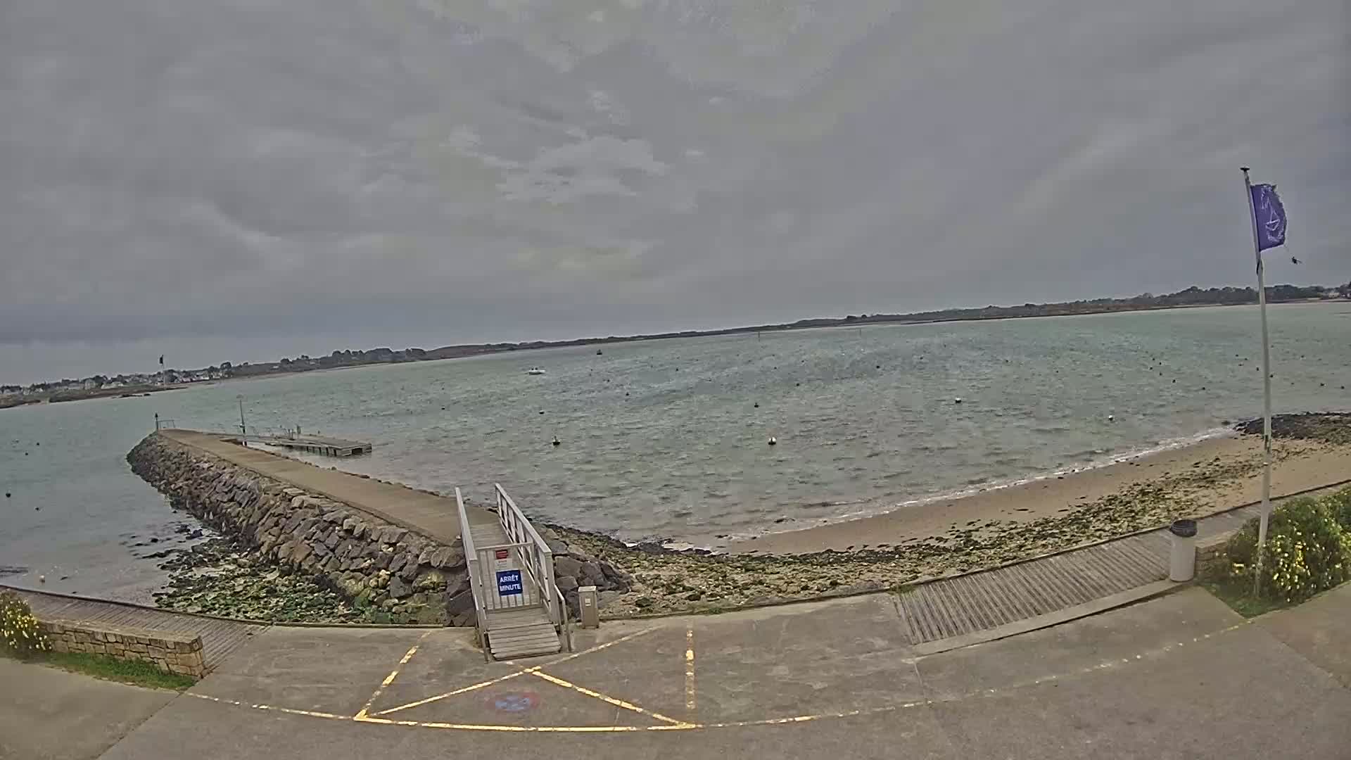 A stone jetty extends into calm, grey water under an overcast sky, leading to a small sandy beach.
