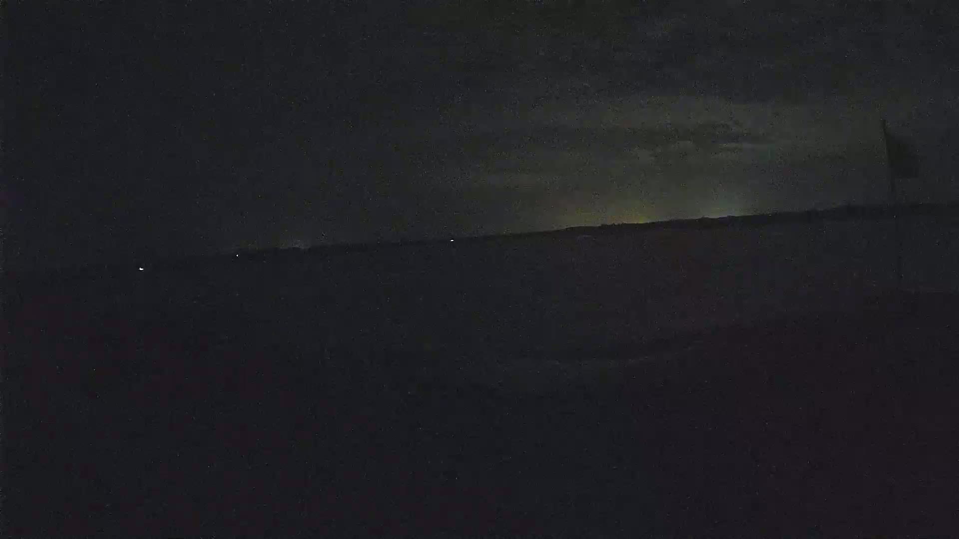 A dark, nighttime landscape shows a faint, distant glow on the horizon under a cloudy sky.