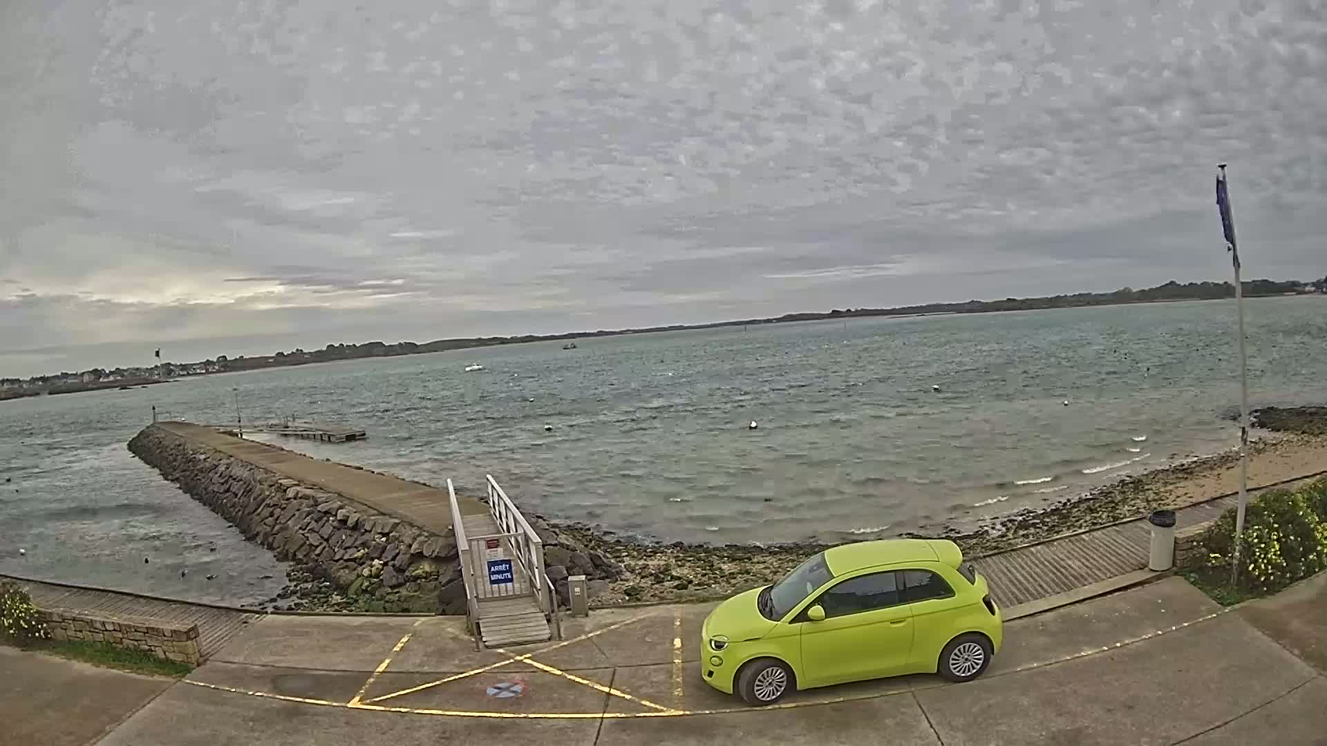 A lime-green car is parked near a stone jetty overlooking a calm, gray-sky day over a body of water.