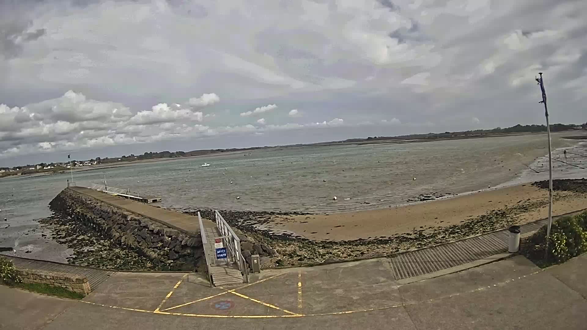 A stone jetty extends into a calm, light-green body of water under a cloudy sky, with a sandy beach visible at low tide.