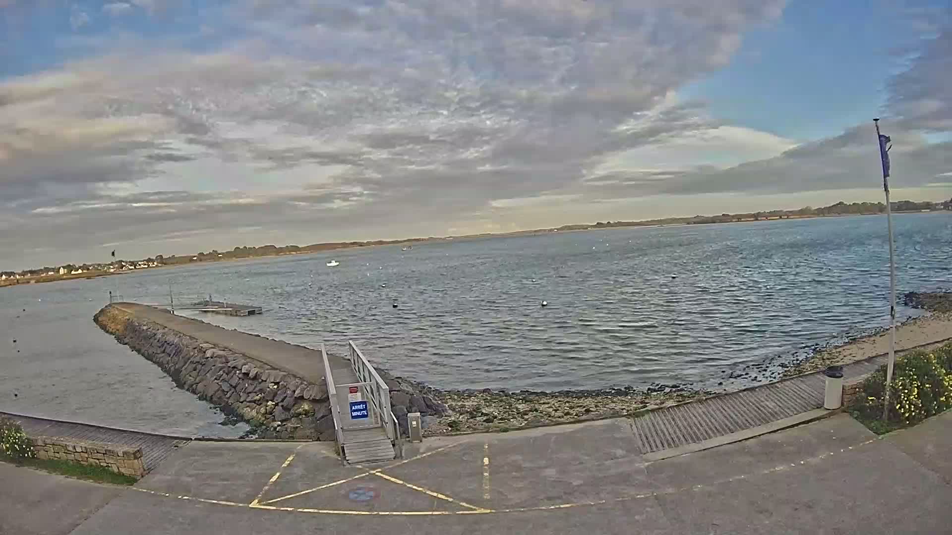 A calm body of water is seen from a paved area leading to a small dock and a stone jetty under a partly cloudy sky.