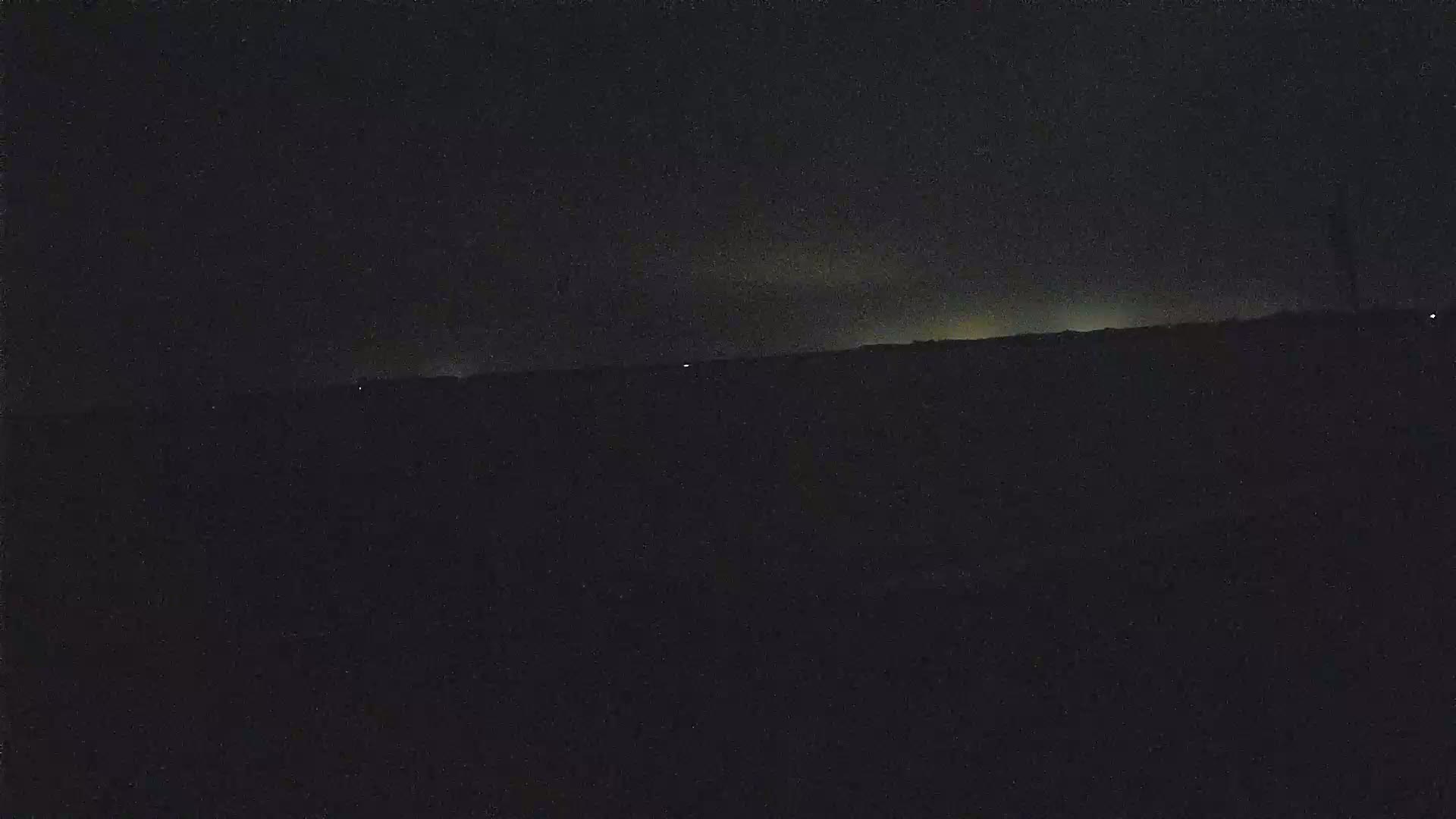A dark, nighttime landscape shows a faintly illuminated horizon under a mostly clear, dark sky.