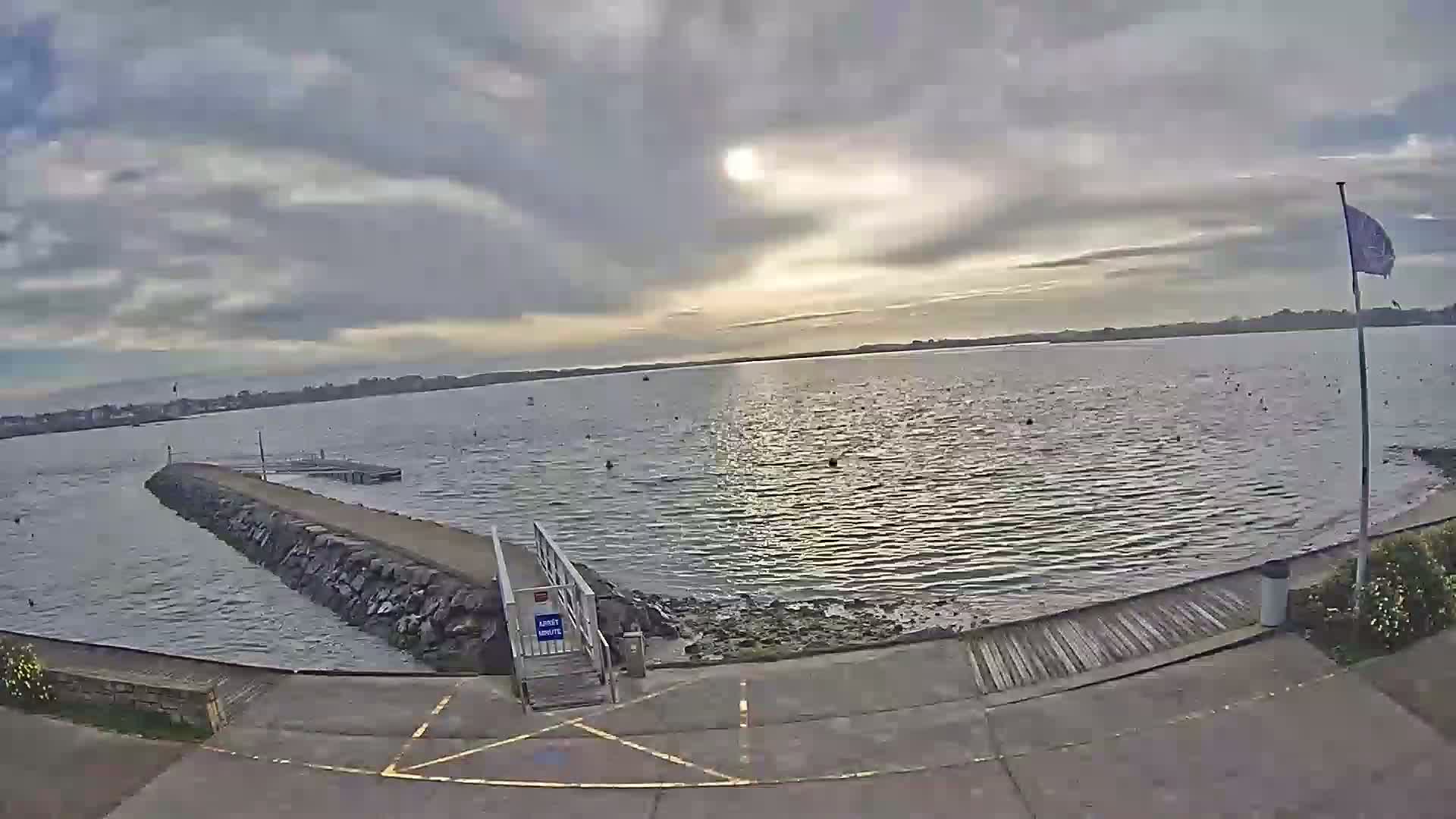 A calm body of water is viewed from a paved area leading to a stone jetty under a mostly cloudy sky.