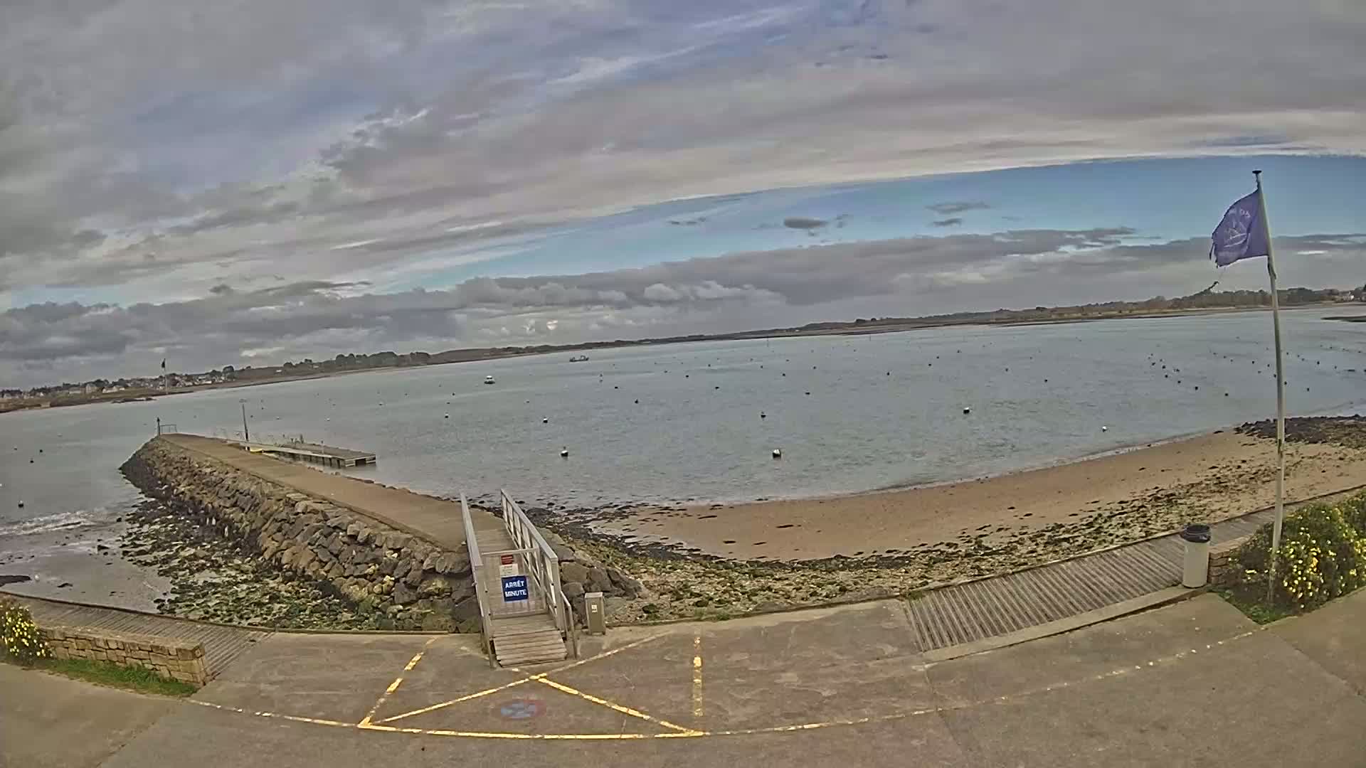 A rocky jetty extends into a calm bay under a partly cloudy sky, with a sandy beach visible to the right.