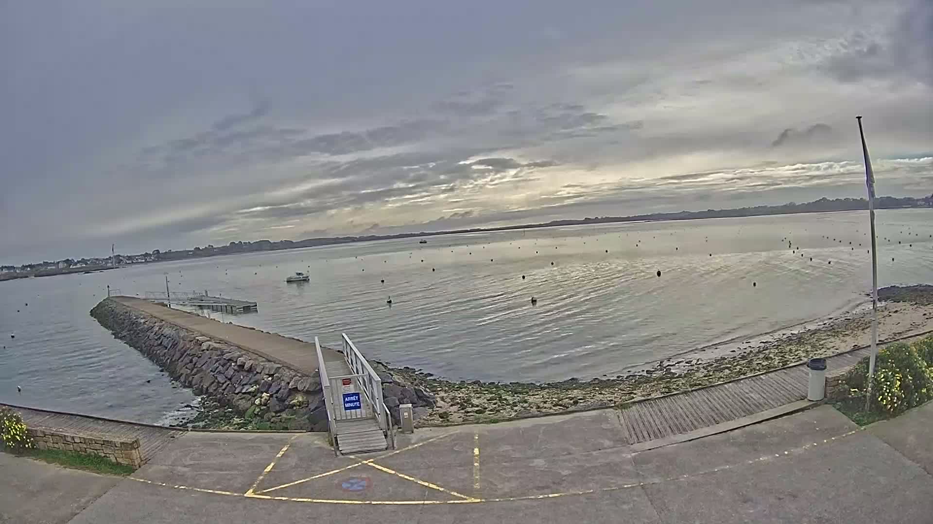 A calm body of water with a stone jetty and a small wooden walkway leading to a ramp, under a mostly cloudy sky.